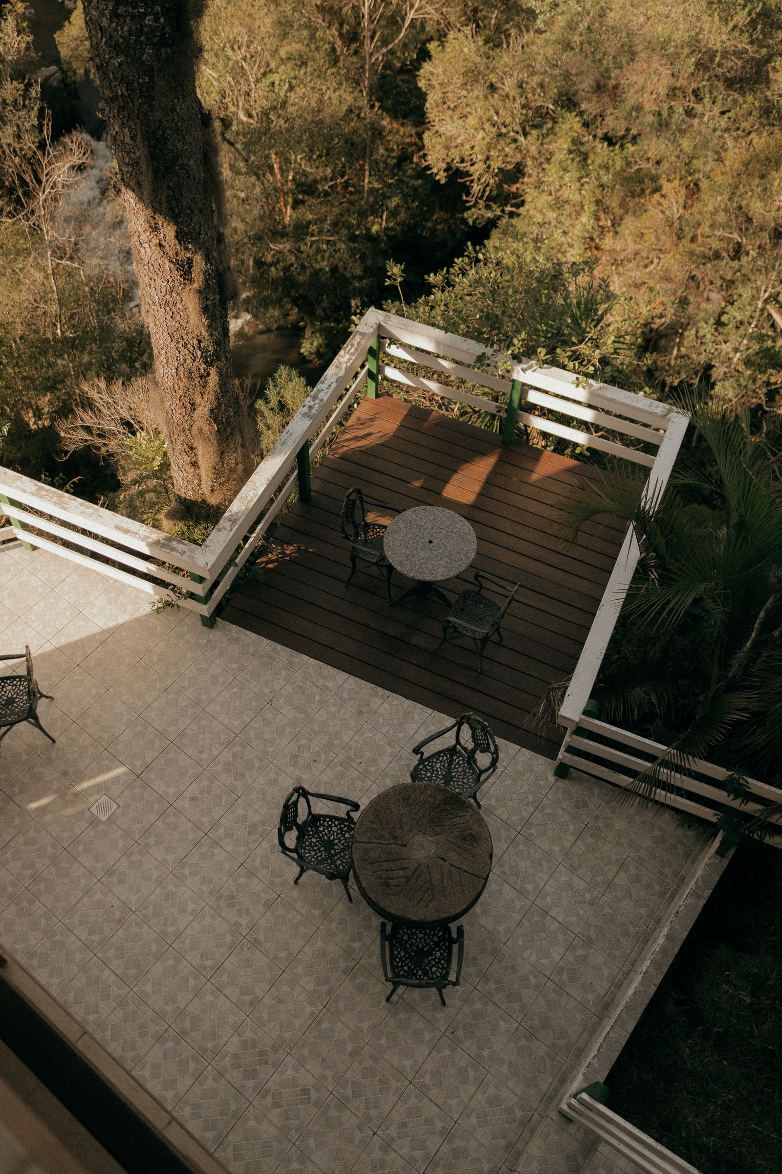 Terrasse en bois avec garde-corps blanc surplombant les arbres, prolongeant un sol carrelé avec tables rondes