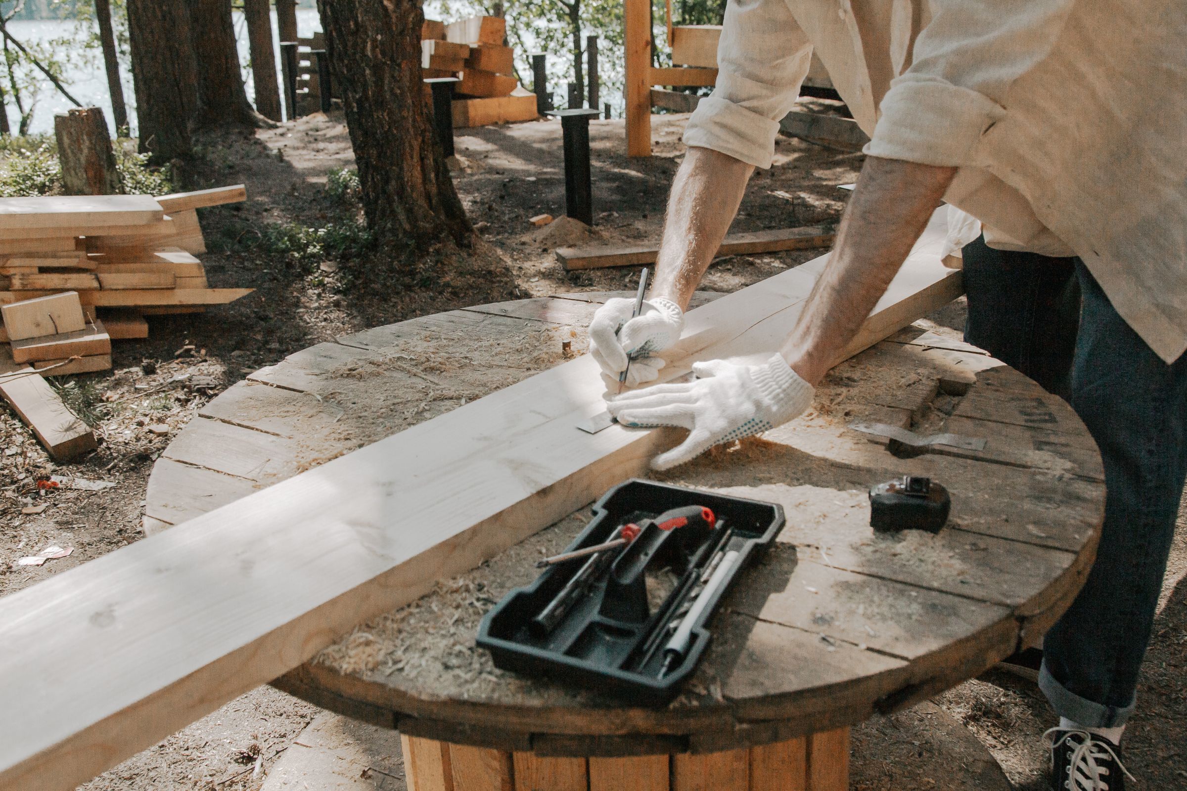 Menuisier traçant au crayon une planche de bois posée sur un touret, avec outils et bois empilé en extérieur