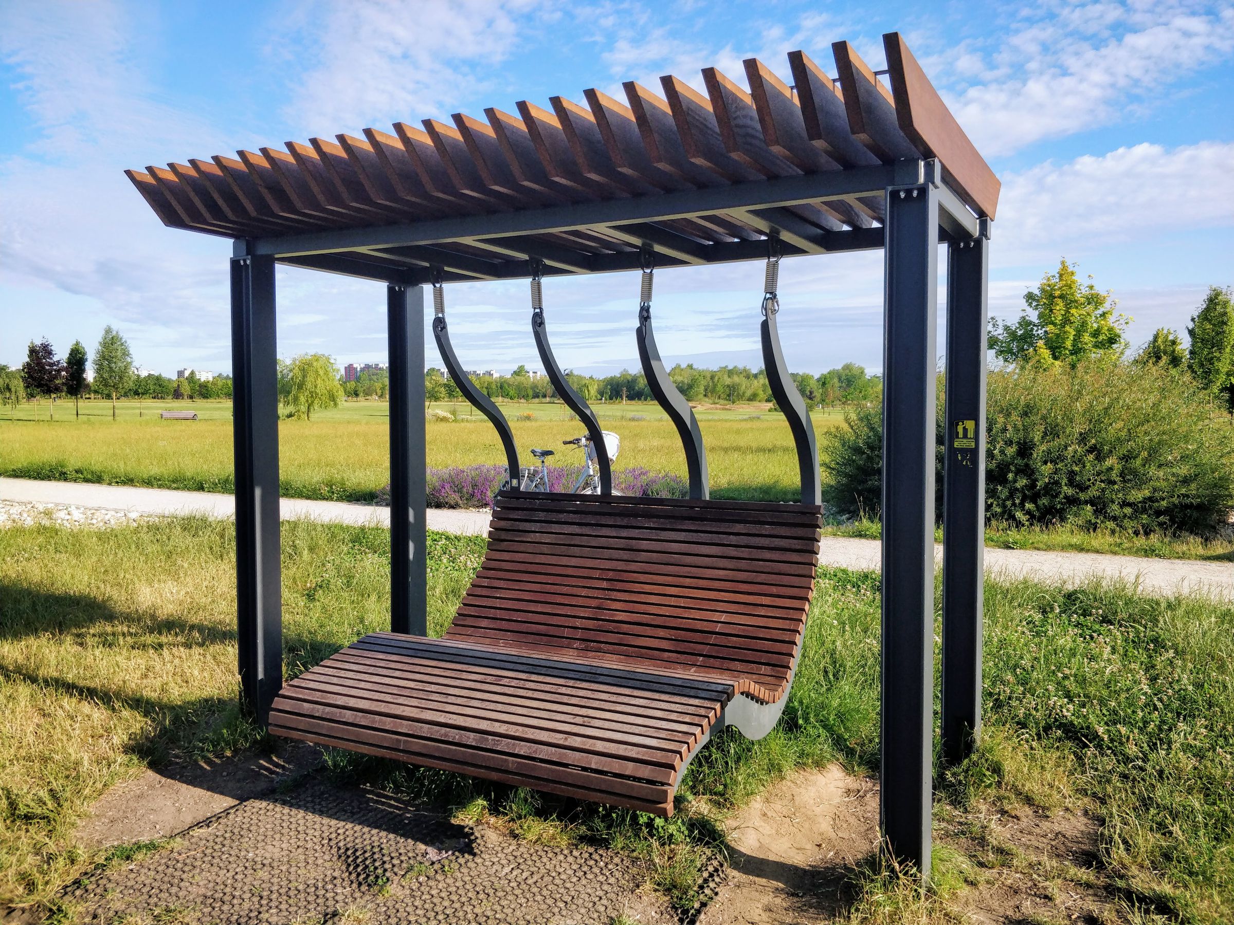Pergola à poteaux en acier et toit de lames de bois abritant une balancelle en bois, au milieu d’une pelouse