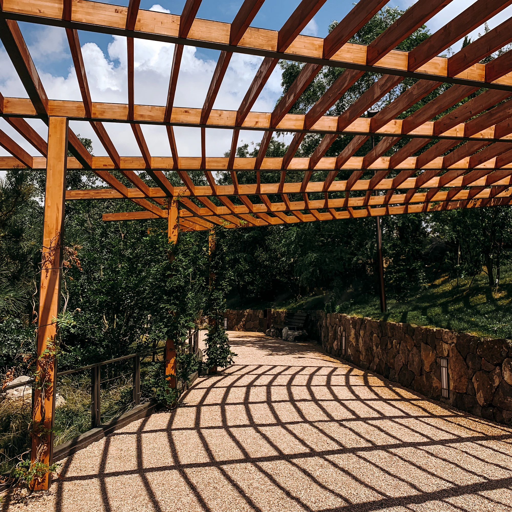 Pergola en bois à toit de lames projetant des ombres rayées sur une allée de gravier bordée d’un mur de pierre