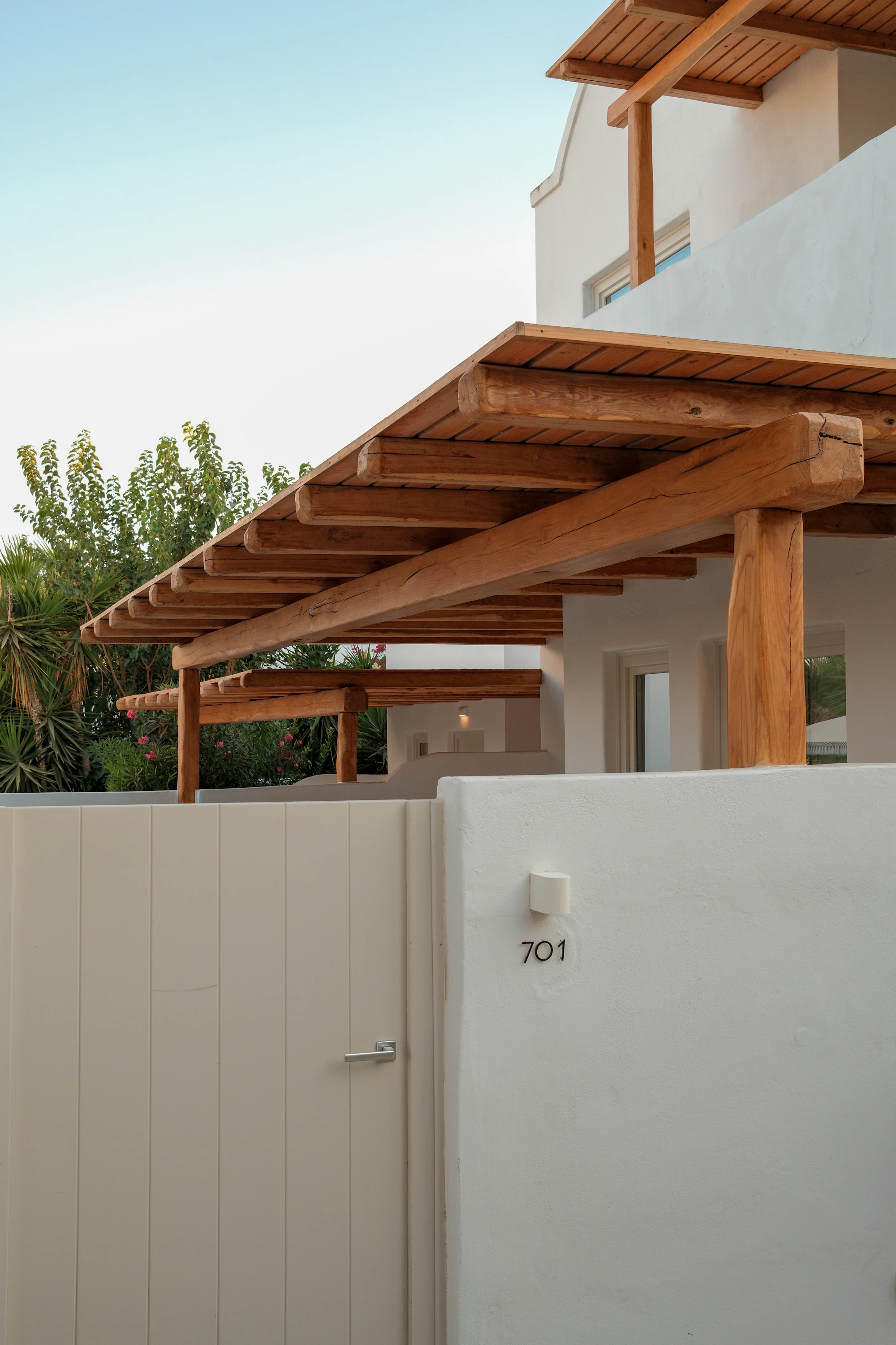 Pergola en bois à poutres brutes et toit de lames, adossée à une maison aux murs blancs près de l’entrée