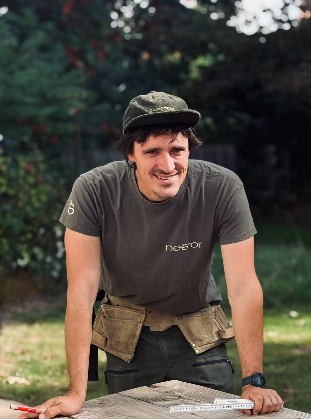 Manu Thirifays, fondateur de Nestor, en t-shirt Nestor et ceinture à outils, accoudé à un plan de travail en bois dans un jardin