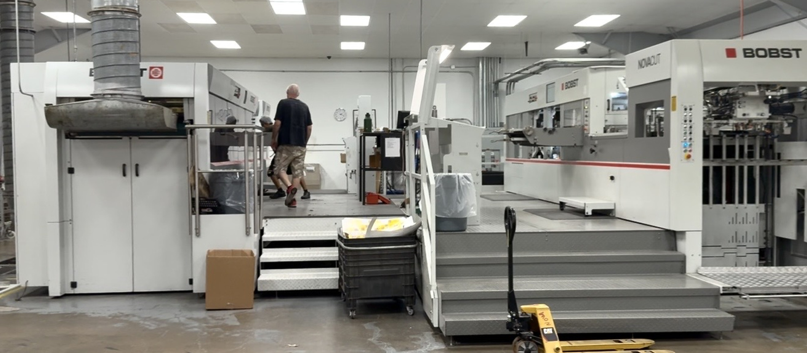 Industrial printing or manufacturing facility with large white and gray Bobst machines on raised platforms, a worker in camo shorts and black shirt walking, and a yellow pallet jack in the foreground on the concrete floor.
