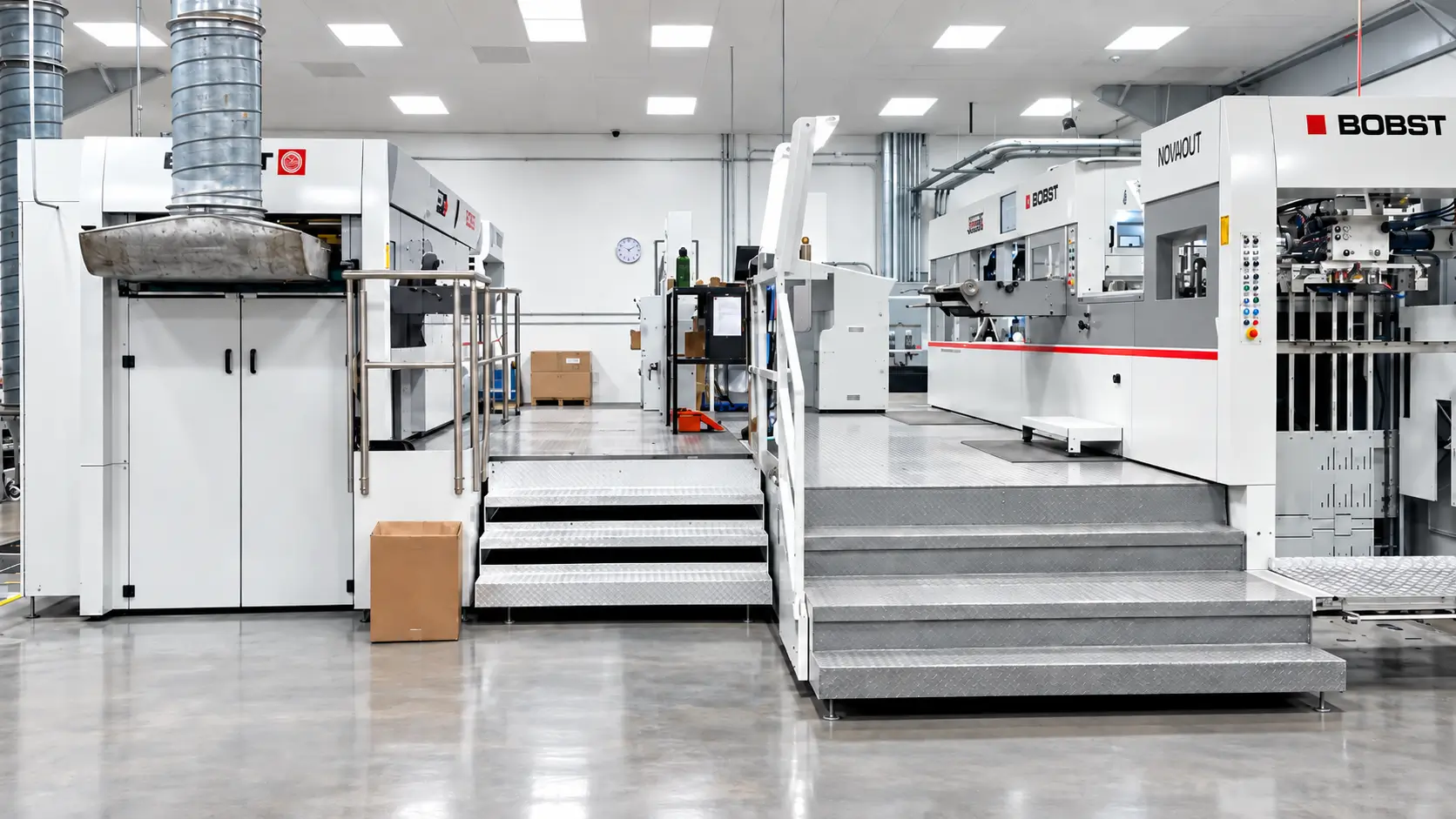 Volara folding carton plant floor with BOBST die-cutters on raised steel platforms and a polished concrete floor.