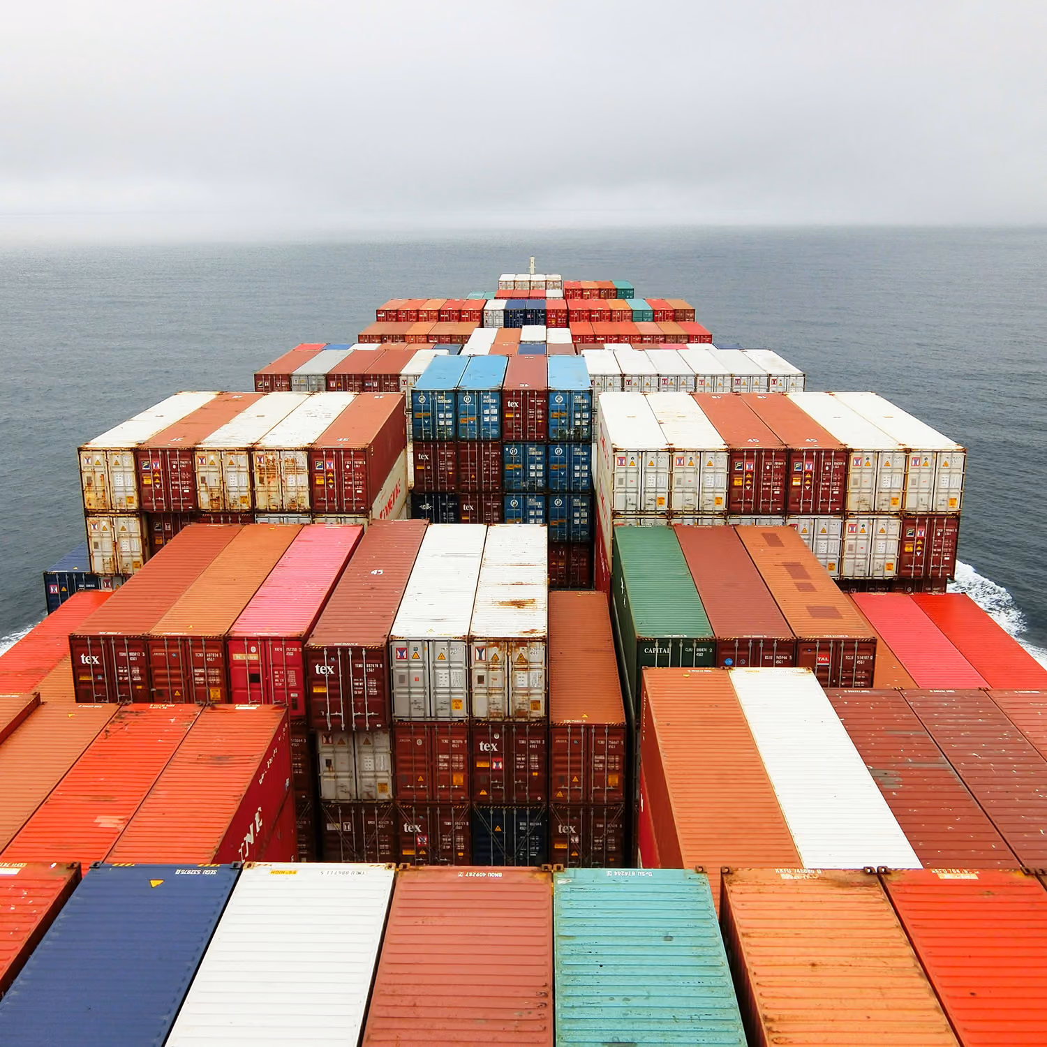 View over stacked multicolored shipping containers on the deck of a cargo ship crossing the ocean under a cloudy sky.