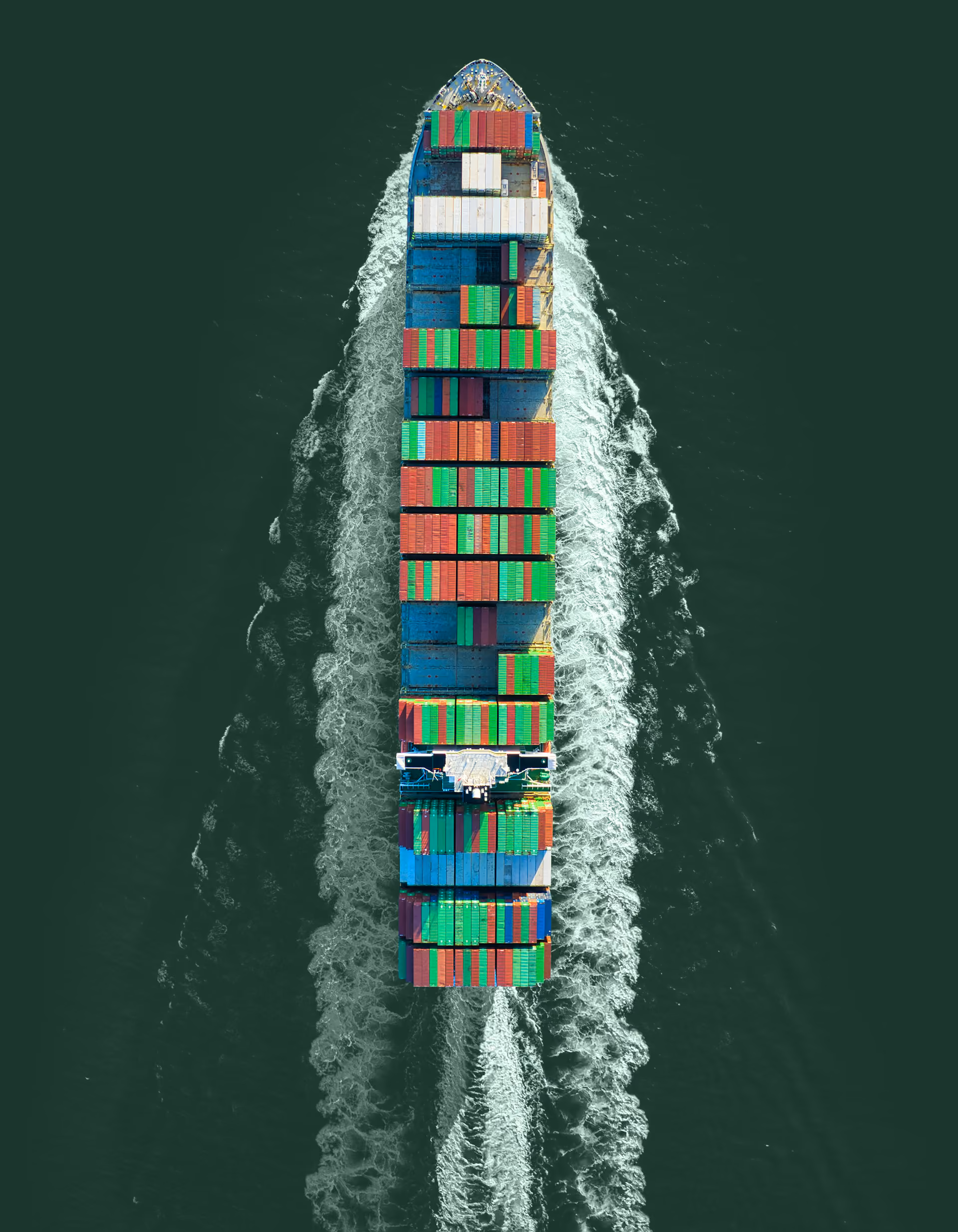 Aerial view of a large cargo ship carrying multicolored shipping containers sailing through deep green water, leaving a white foamy wake behind.