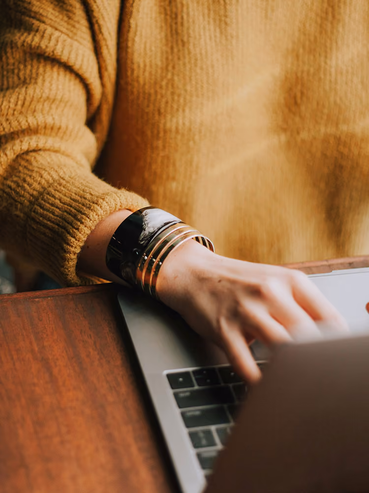 Person wearing a mustard yellow knit sweater typing on a laptop keyboard with a black and gold bracelet on their wrist.