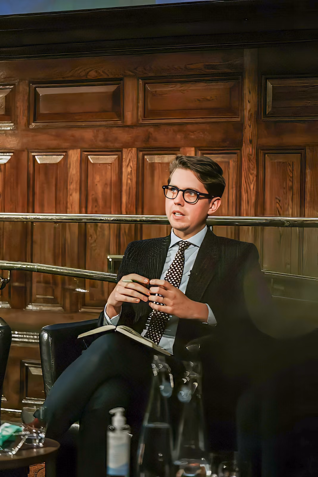 Young man in a dark suit and patterned tie wearing glasses, sitting on a leather chair in a wood-paneled room, speaking with hands clasped and an open notebook on his lap.