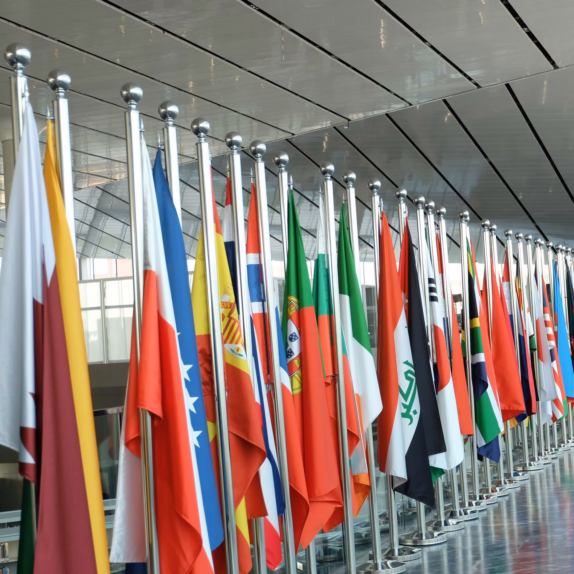 Row of various national flags displayed on silver poles inside a modern building with a reflective ceiling and glass walls.
