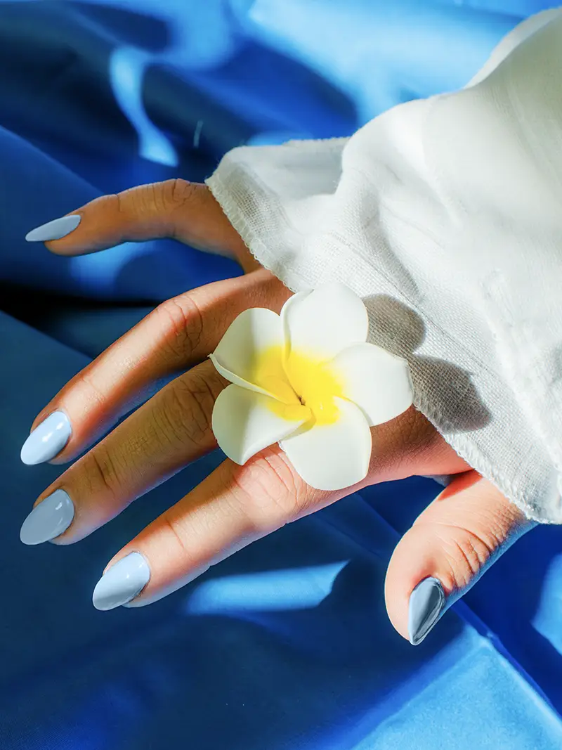 Photo of hands with light blue painted nails, holding a white and yellow flower, with a blue backdrop