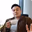 A man with short black hair wearing a brown jacket over a gray shirt, holding a small white cup and gesturing with his other hand, looking slightly upward.
