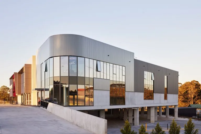 Modern commercial building with large floor-to-ceiling glass windows, a curved corner, and a combination of grey metal paneling and concrete base, surrounded by a parking area and landscaped greenery under a clear sky.