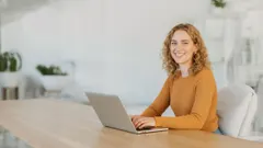 A woman with curly hair wearing an orange sweater sitting at a wooden table and typing on a laptop, smiling at the camera in a bright, modern room.