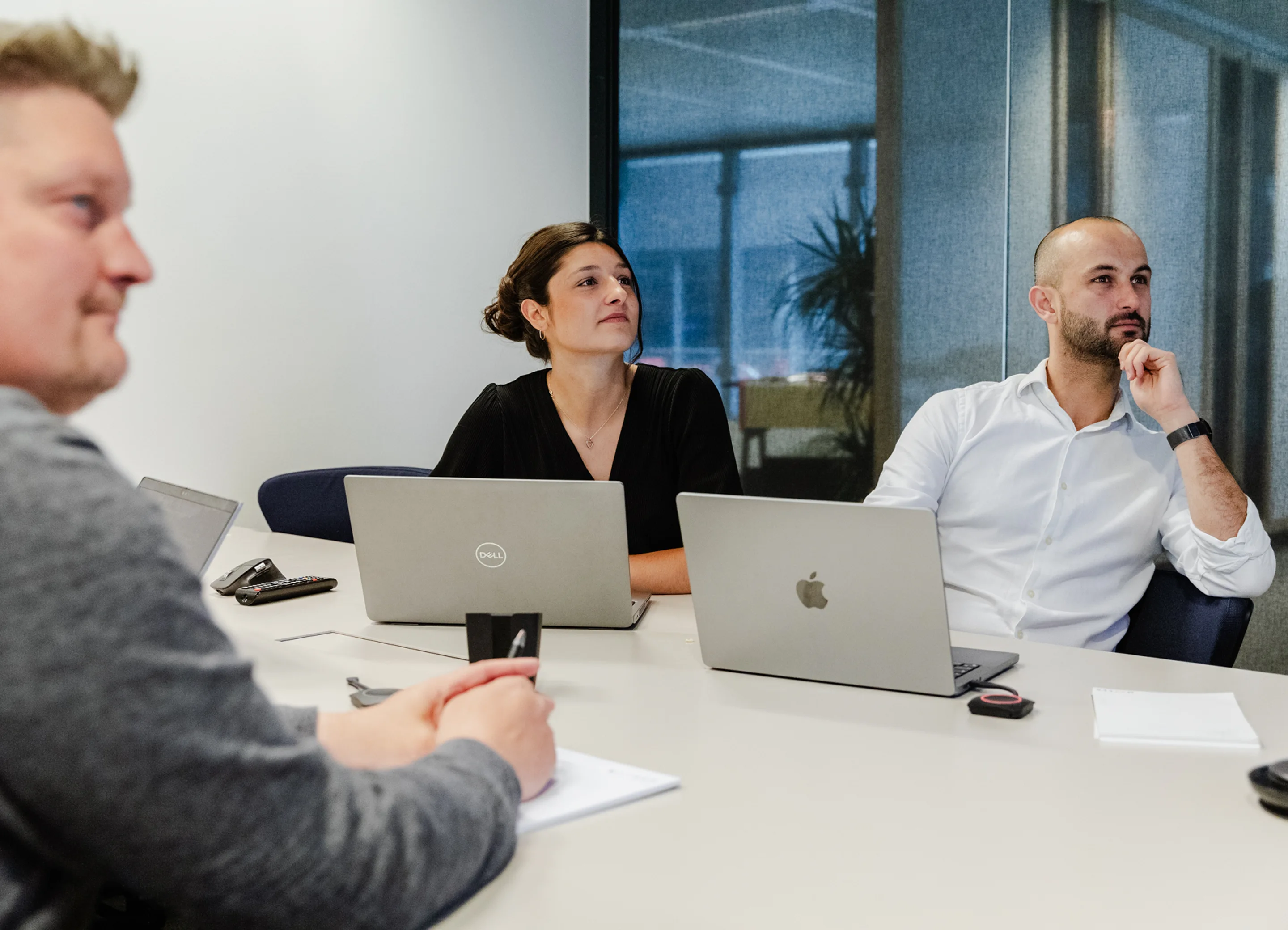 Three people sitting at a conference table with laptops and notebooks during a meeting, attentively looking toward something outside the frame.