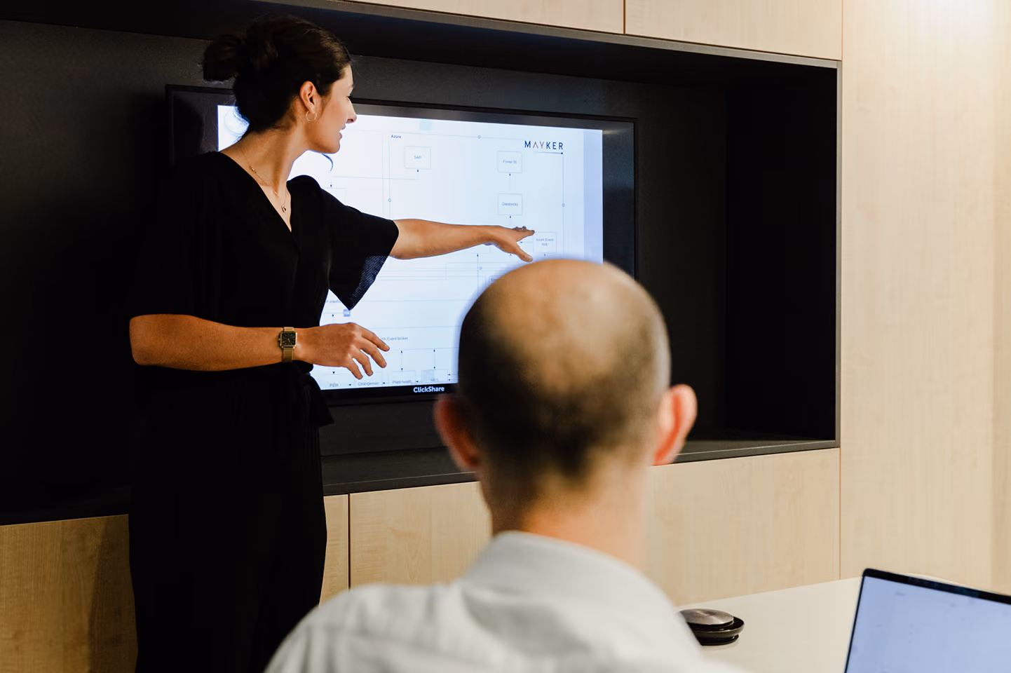 A woman in black points at a screen displaying a presentation while a man with a shaved head watches, seated at a table with a laptop.