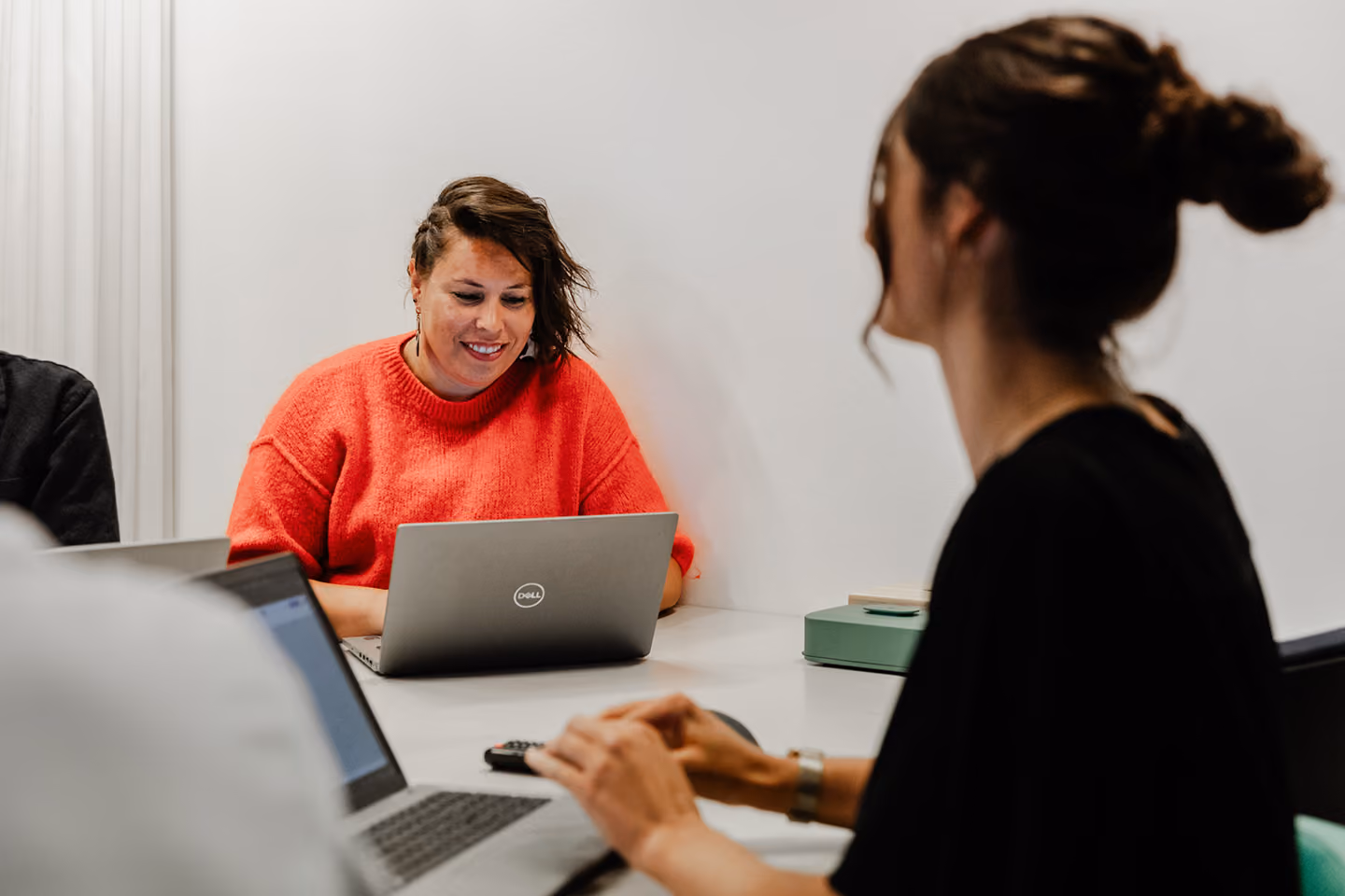 A woman wearing a bright red sweater smiles while working on a Dell laptop at a table with others using laptops in a bright room.