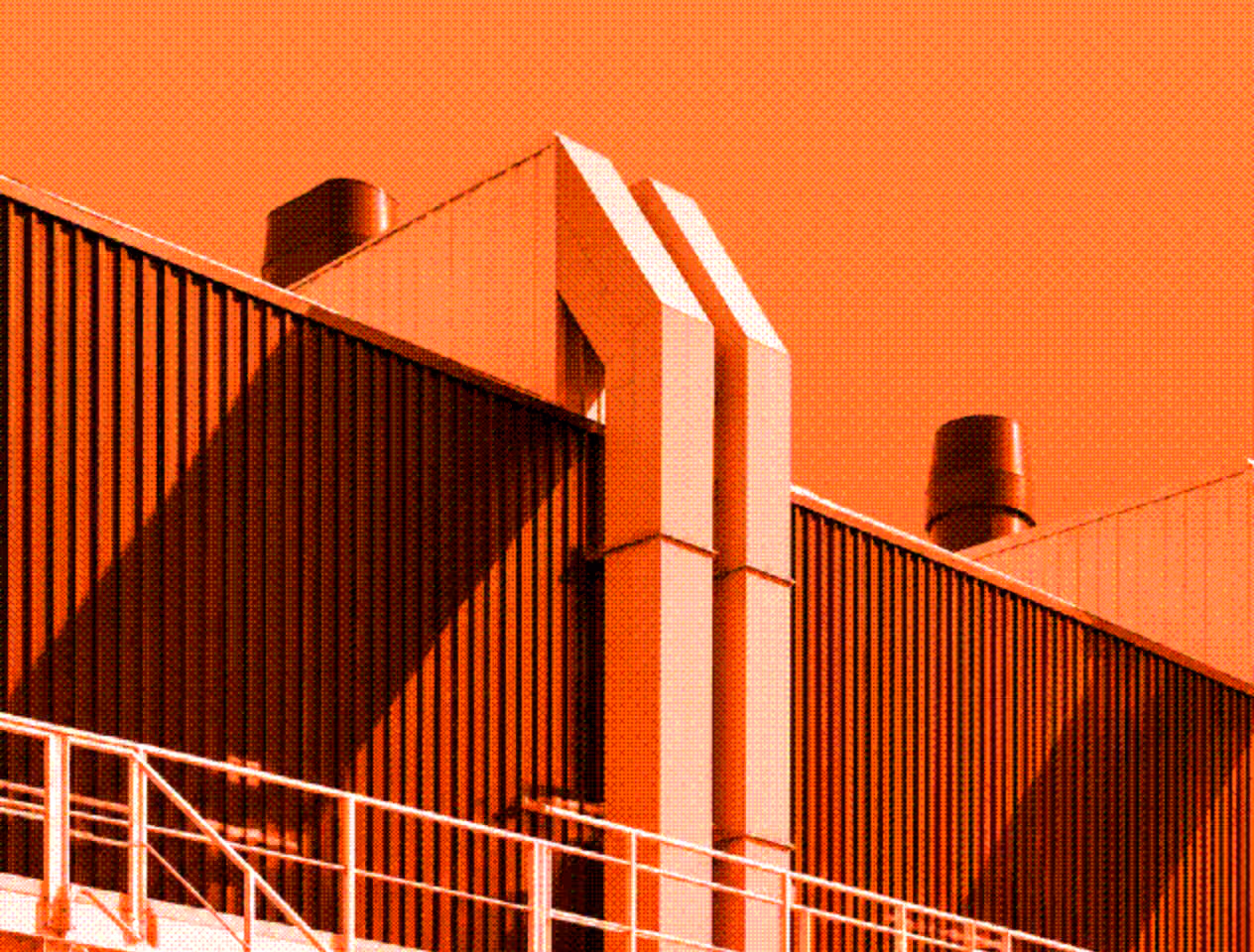 Close-up of an industrial building with large vertical metal panels, two prominent vertical chimneys or exhaust pipes, and metal railings on an exterior platform against an orange sky.