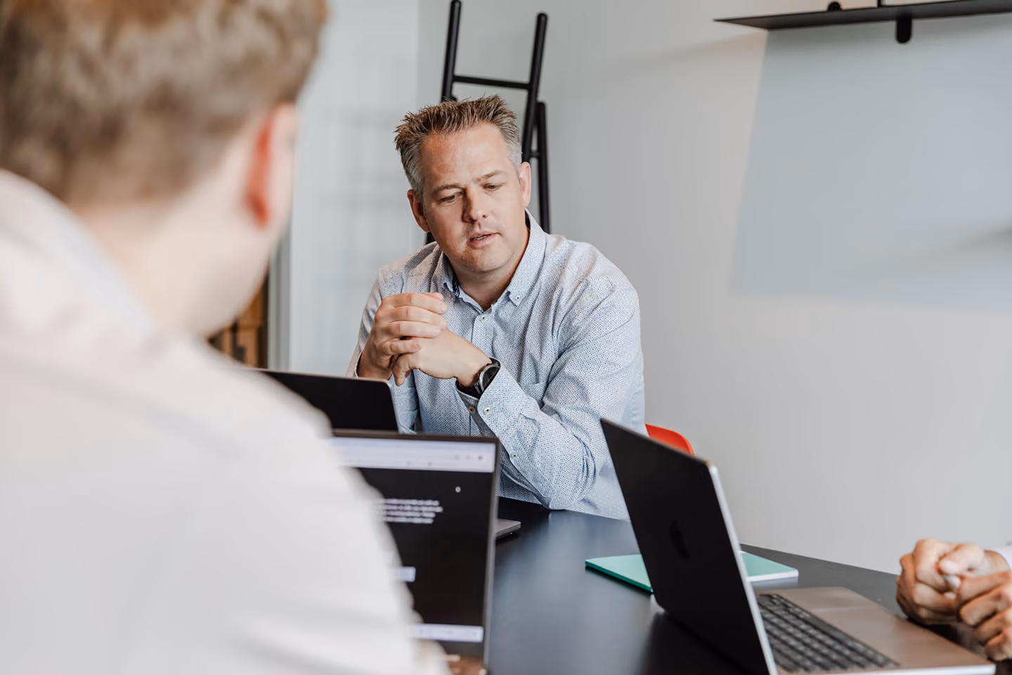 Man in a light blue shirt sitting at a table with laptops, appearing to explain or discuss something with two people partially visible across from him.