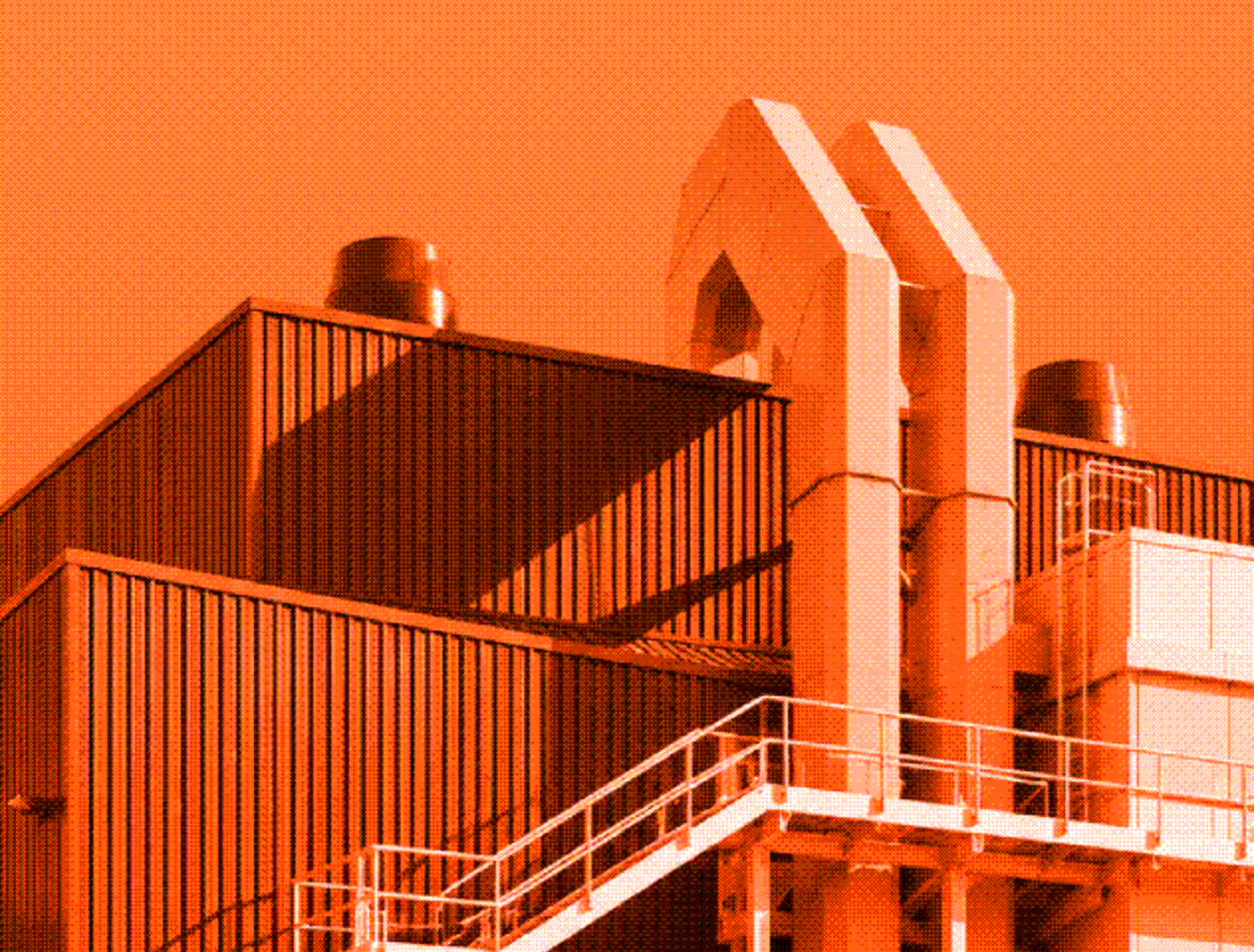 Close-up view of industrial building sections with metal siding, pipes, chimney vents, and external staircase under an orange sky.