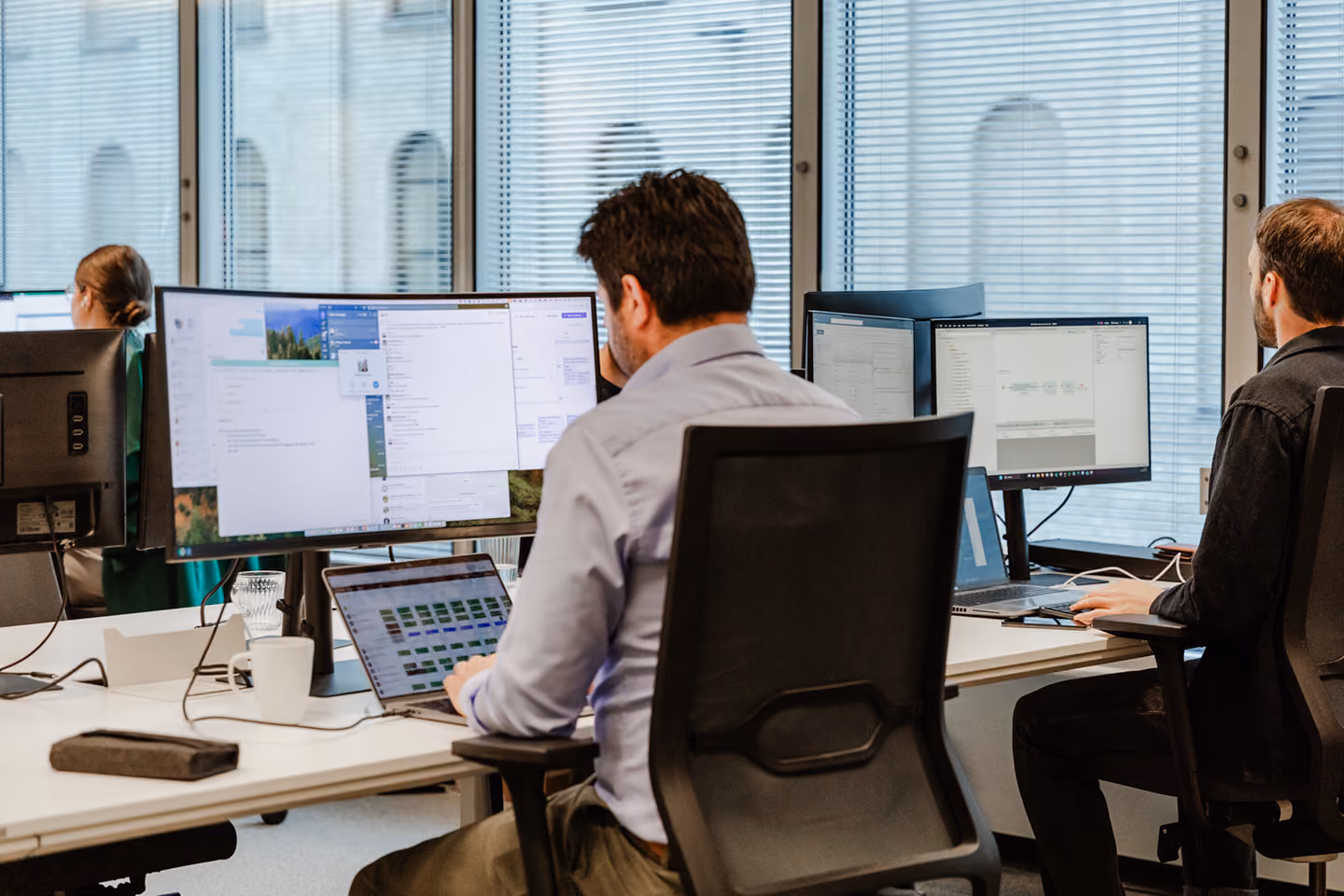 Two men seated at a shared office desk, each working on desktop computers with multiple monitors, near large windows with blinds.