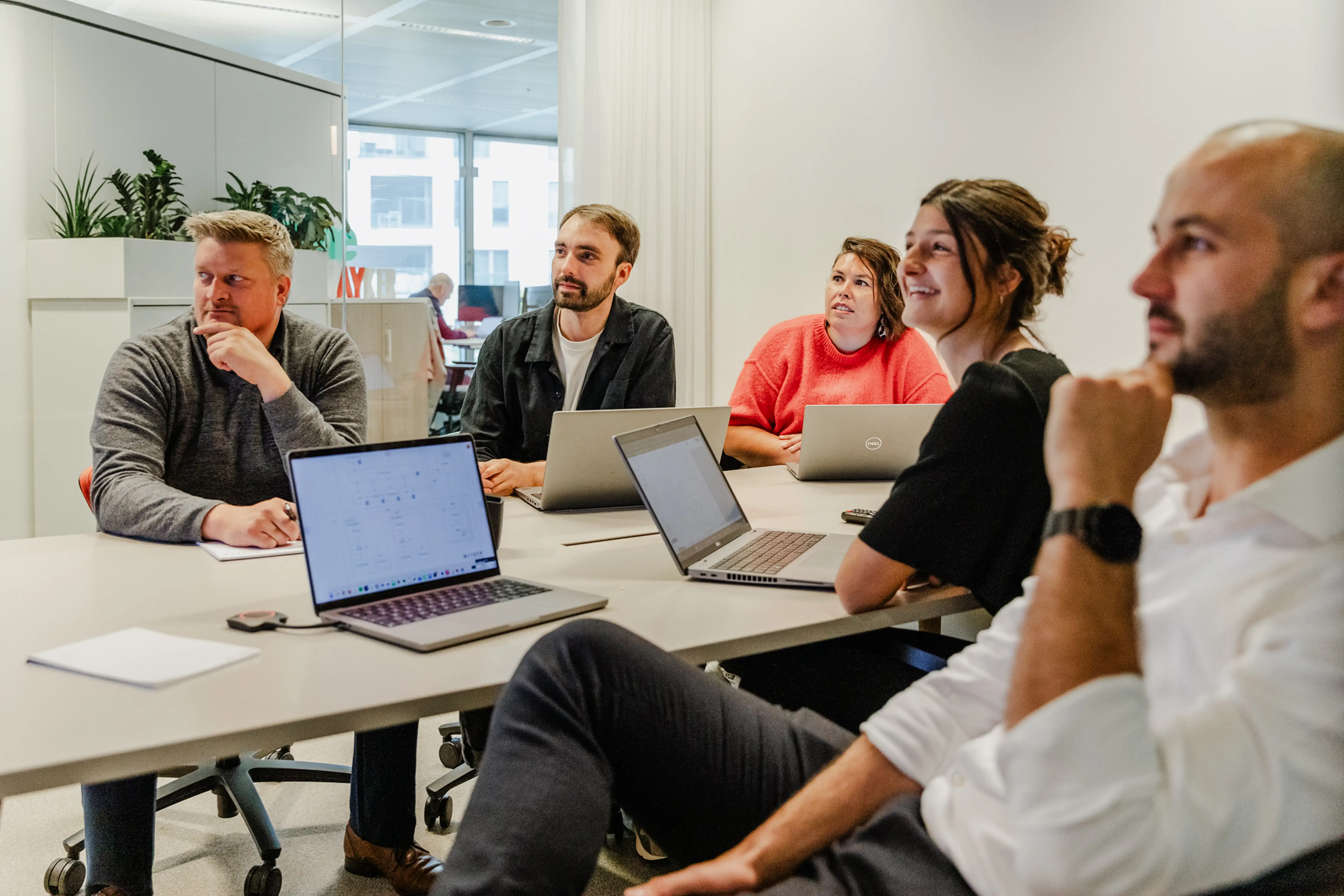 Five people sitting around a conference table with laptops, engaged in a meeting or discussion in a modern office with white walls and large windows.
