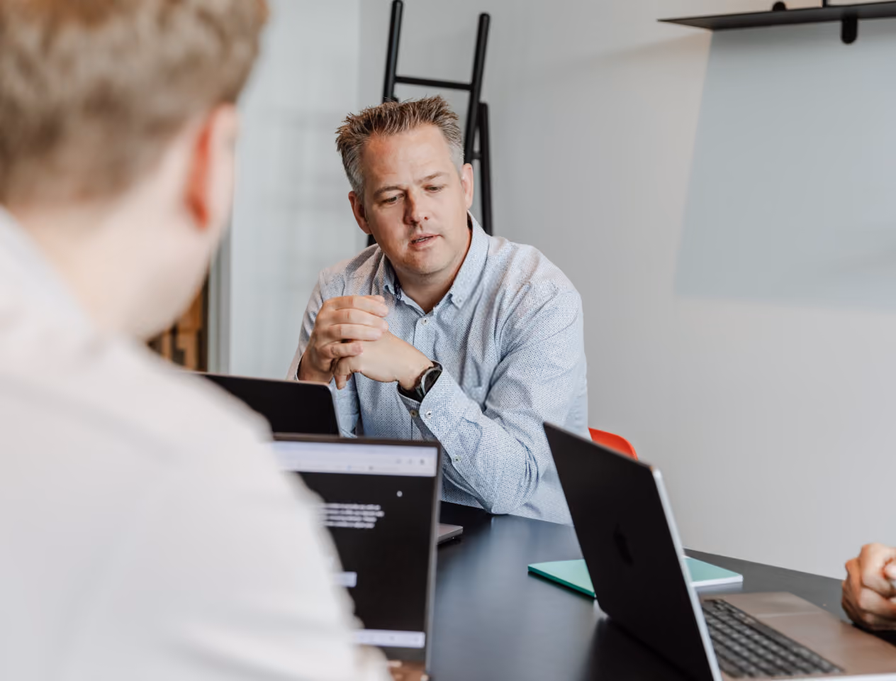 Man in a light blue shirt speaking or explaining something in a meeting with others around a table with laptops.