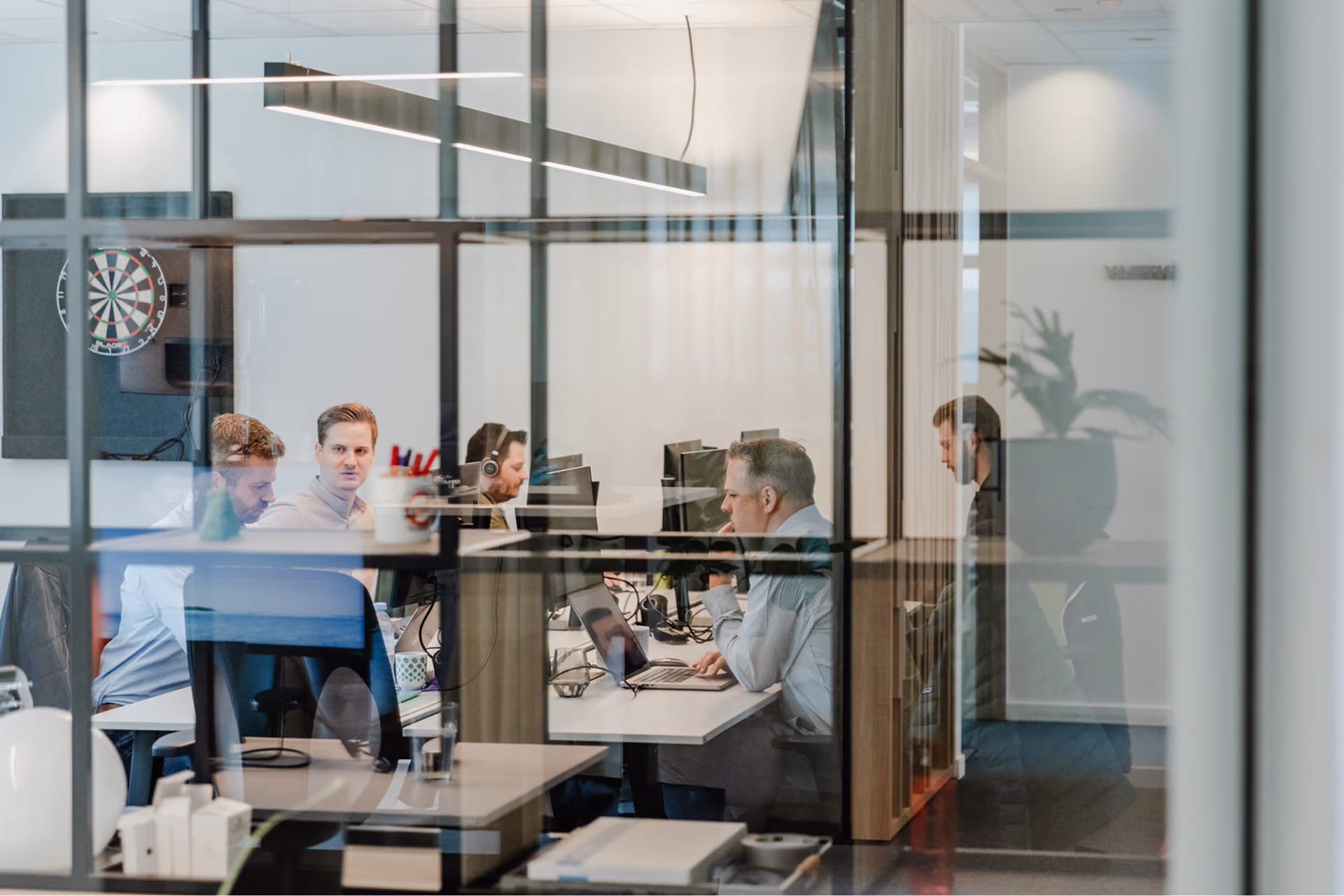 Office scene viewed through glass panels showing five men working at desks with computers in a modern workspace, including one person wearing headphones and another looking at a laptop.