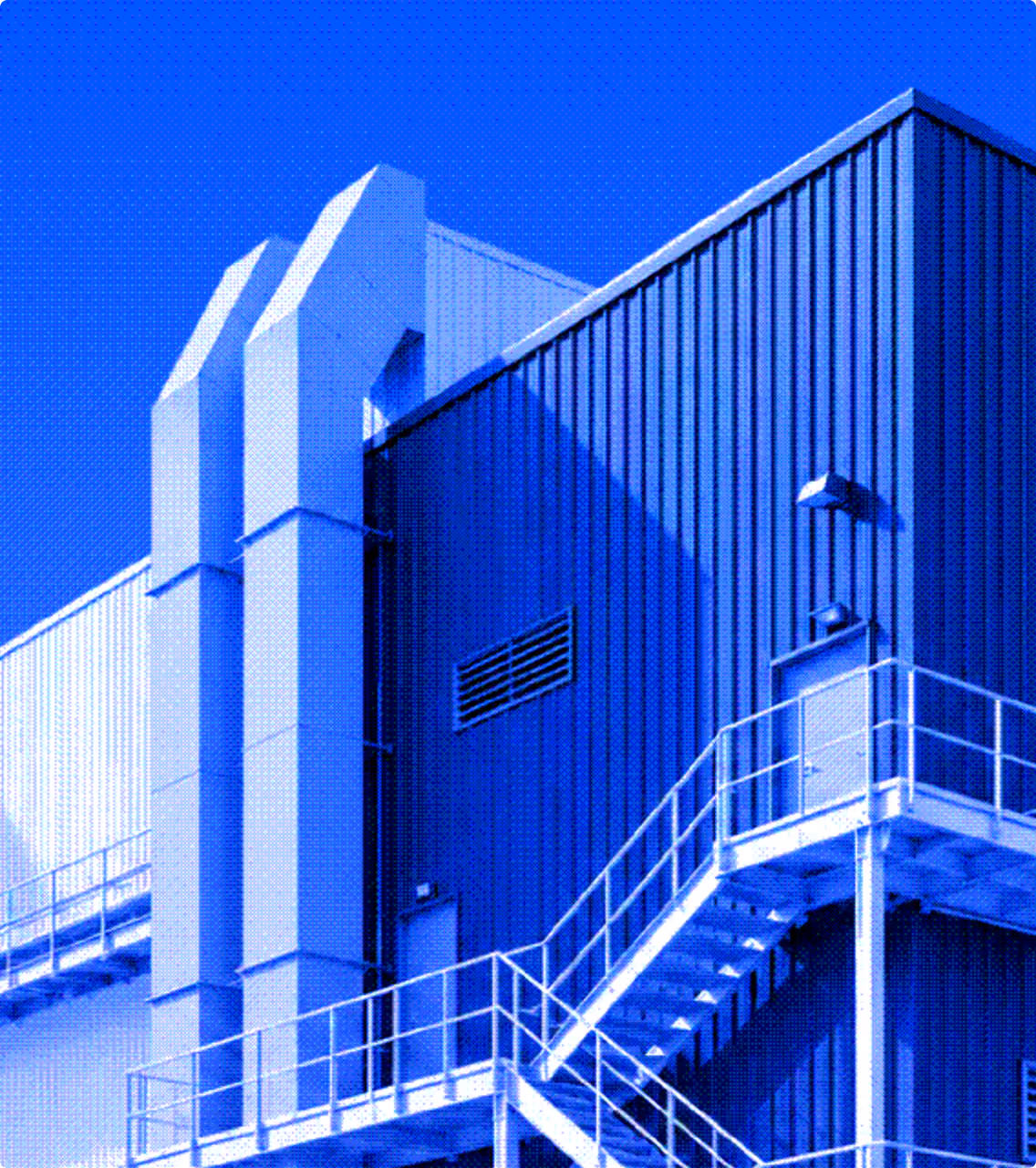 Exterior of a modern industrial building with blue corrugated metal walls, white metal stairs and railings leading to a door, and large white ventilation ducts against a clear bright blue sky.