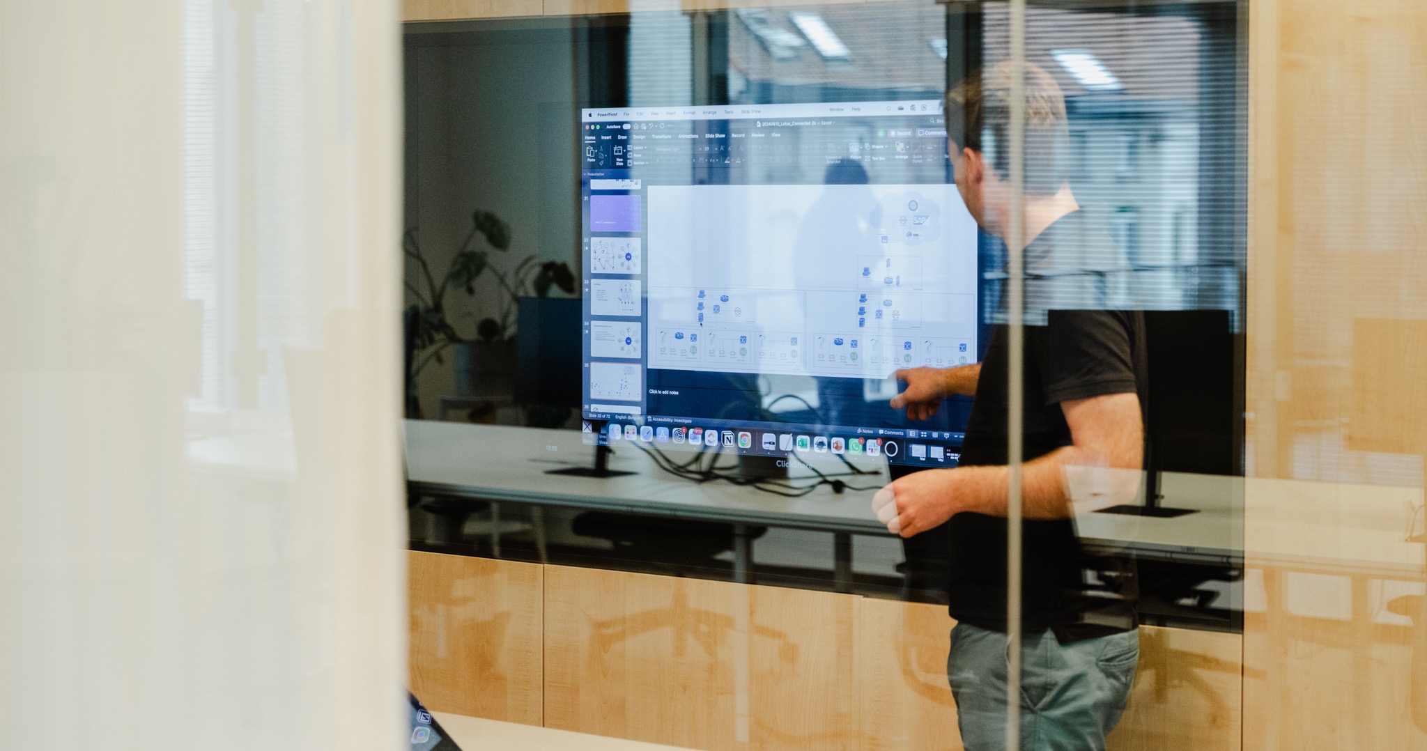 Mayker engineer pointing at a system architecture diagram on a large screen, seen through a glass wall.