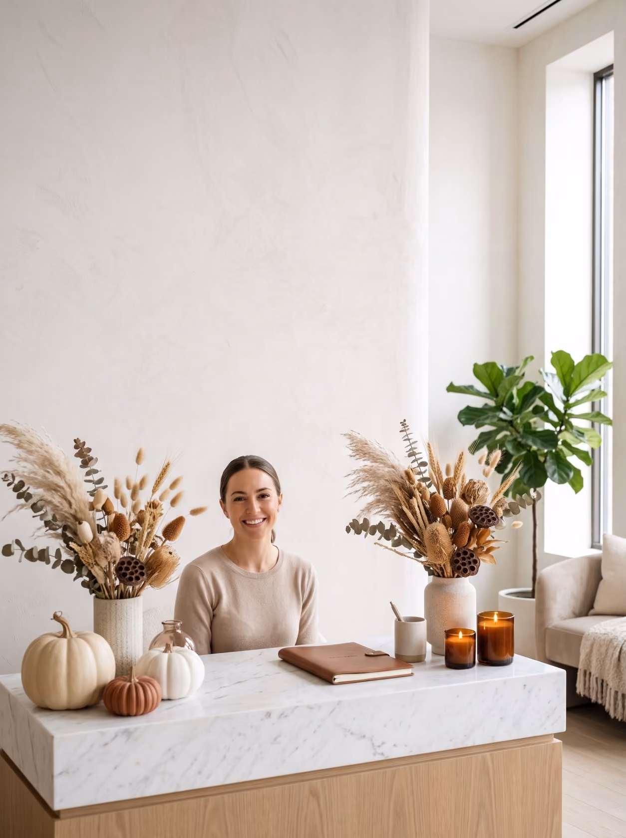 Smiling woman seated behind a white marble desk with neutral decorative dried flower arrangements, pumpkins, candles, and a notebook, in a bright, modern room with large windows and a green potted plant.