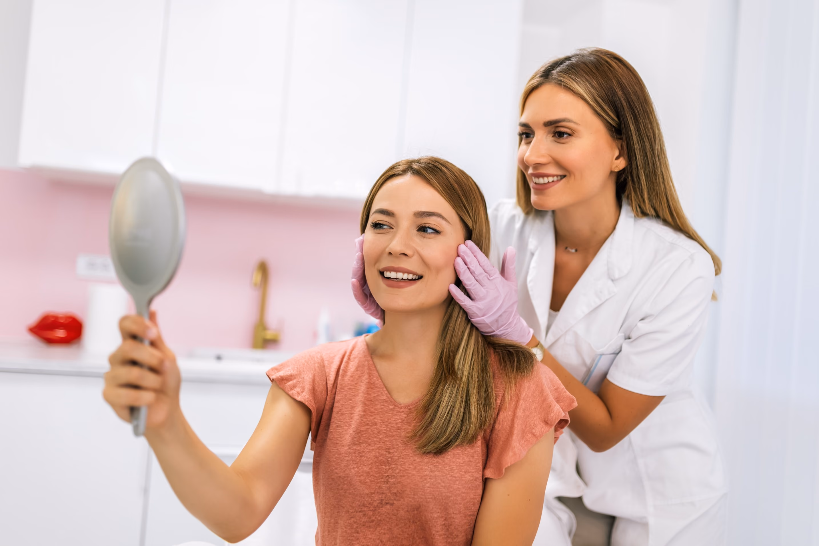 A woman in a coral shirt holds a handheld mirror and smiles at her reflection while a female dermatologist in a white coat and purple gloves examines her face in a clinical setting.