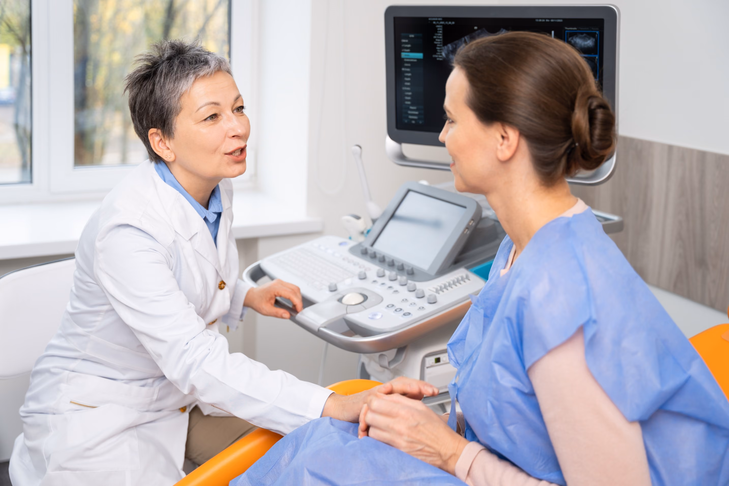A female doctor in a white coat holding hands and speaking with a female patient wearing a blue medical gown in a clinical room with ultrasound equipment nearby.