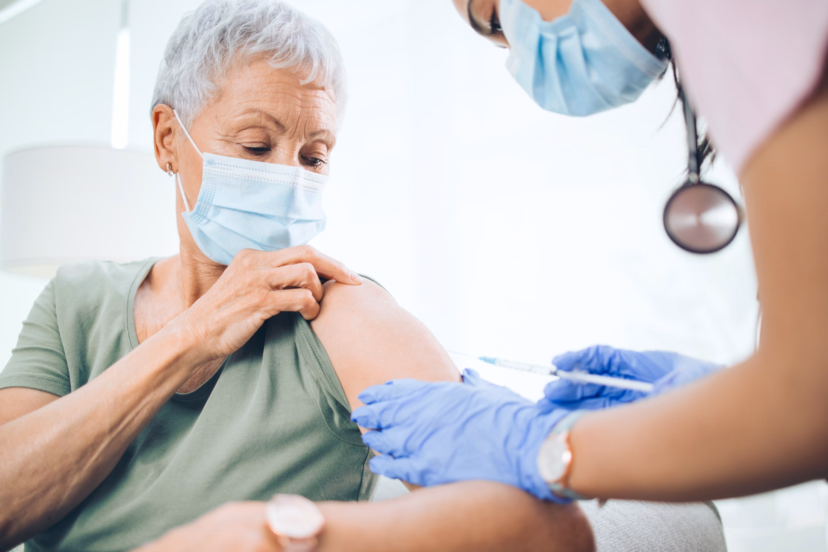 Senior woman wearing a medical mask receiving a vaccine injection in her upper arm from a healthcare worker wearing gloves and a mask.