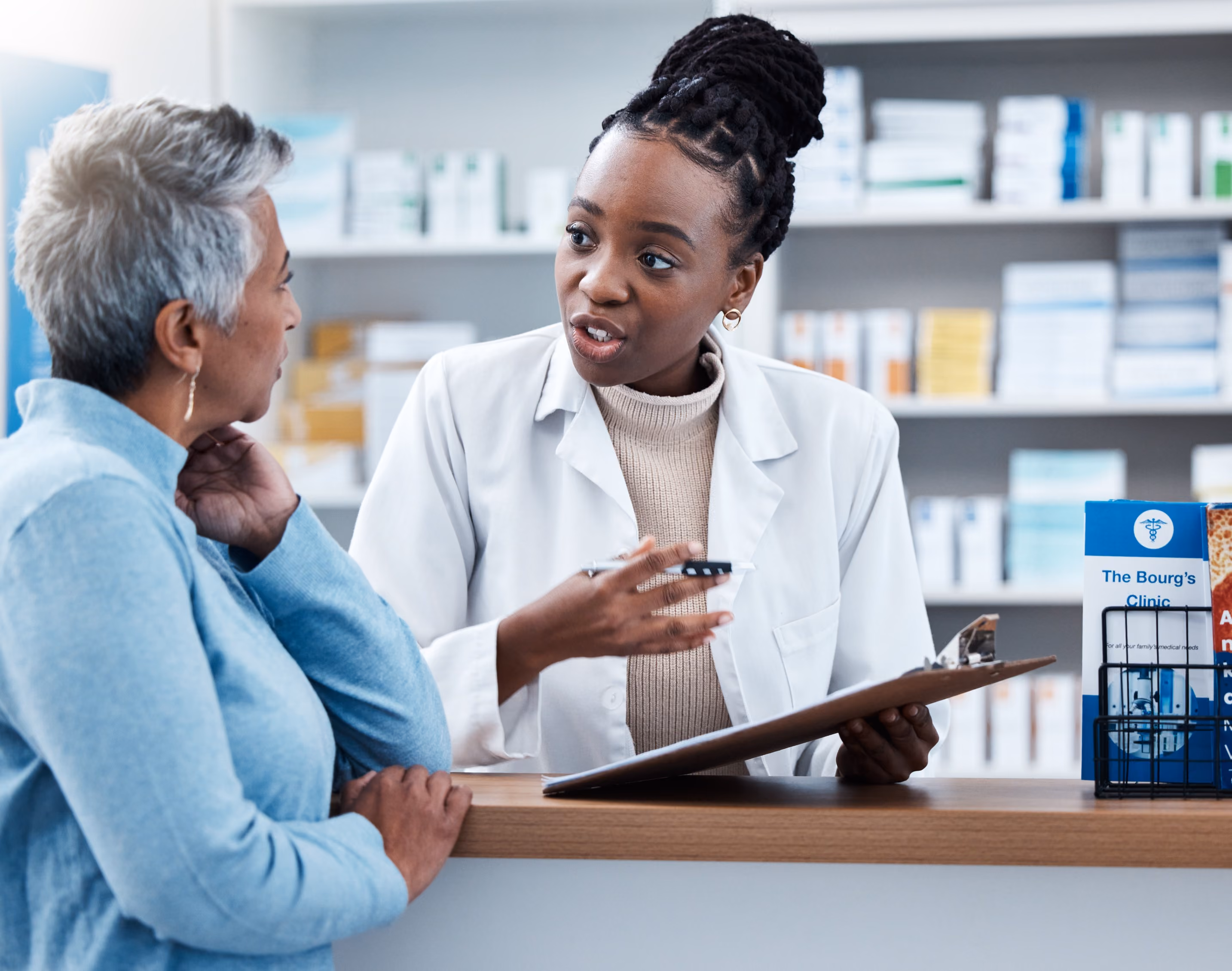 A female pharmacist in a white coat talks to an older woman at a pharmacy counter, holding a clipboard and explaining medication information while the woman listens attentively.