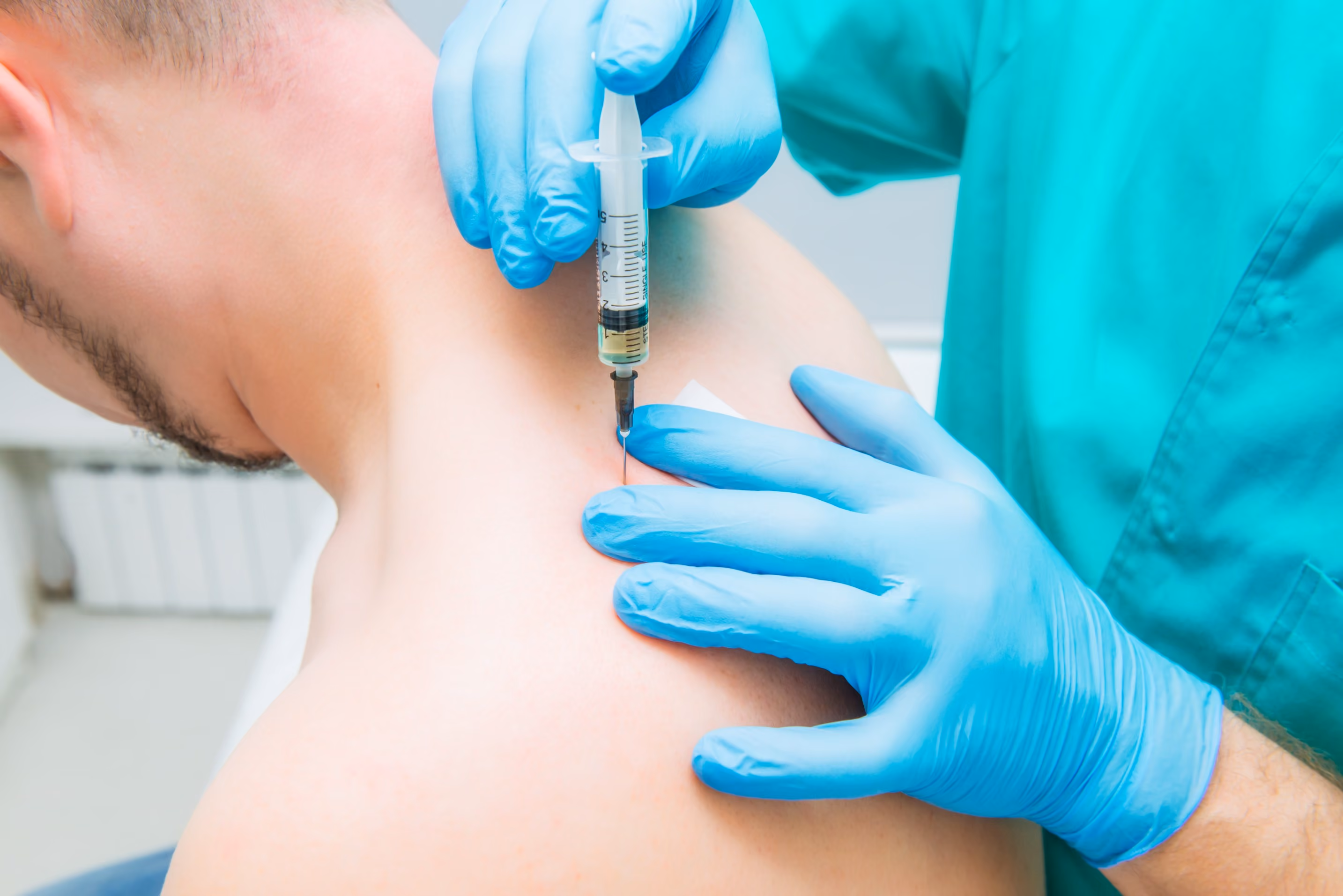A person receiving an injection in the upper back from a healthcare professional wearing blue gloves and a teal uniform.