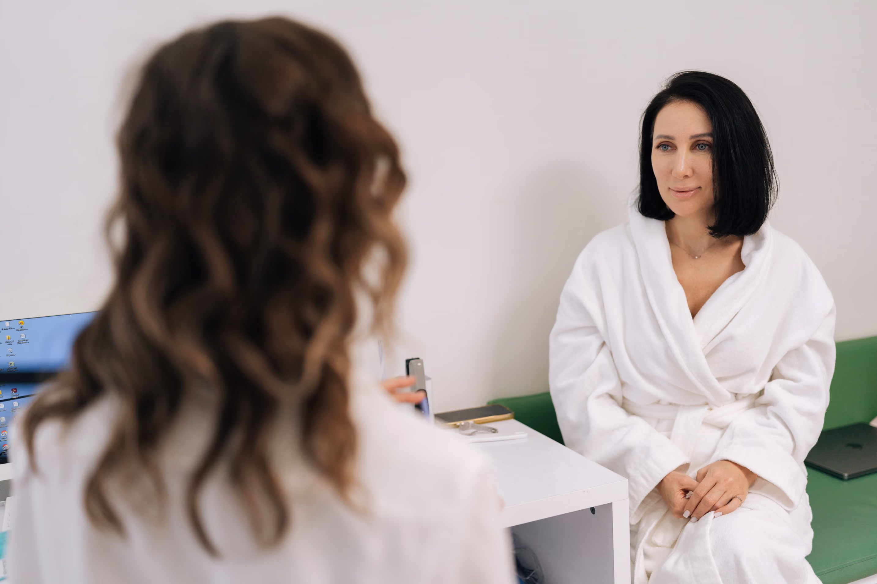 A woman with straight black hair wearing a white robe sits on a green bench facing another woman with curly brown hair, both engaged in conversation in a bright room.