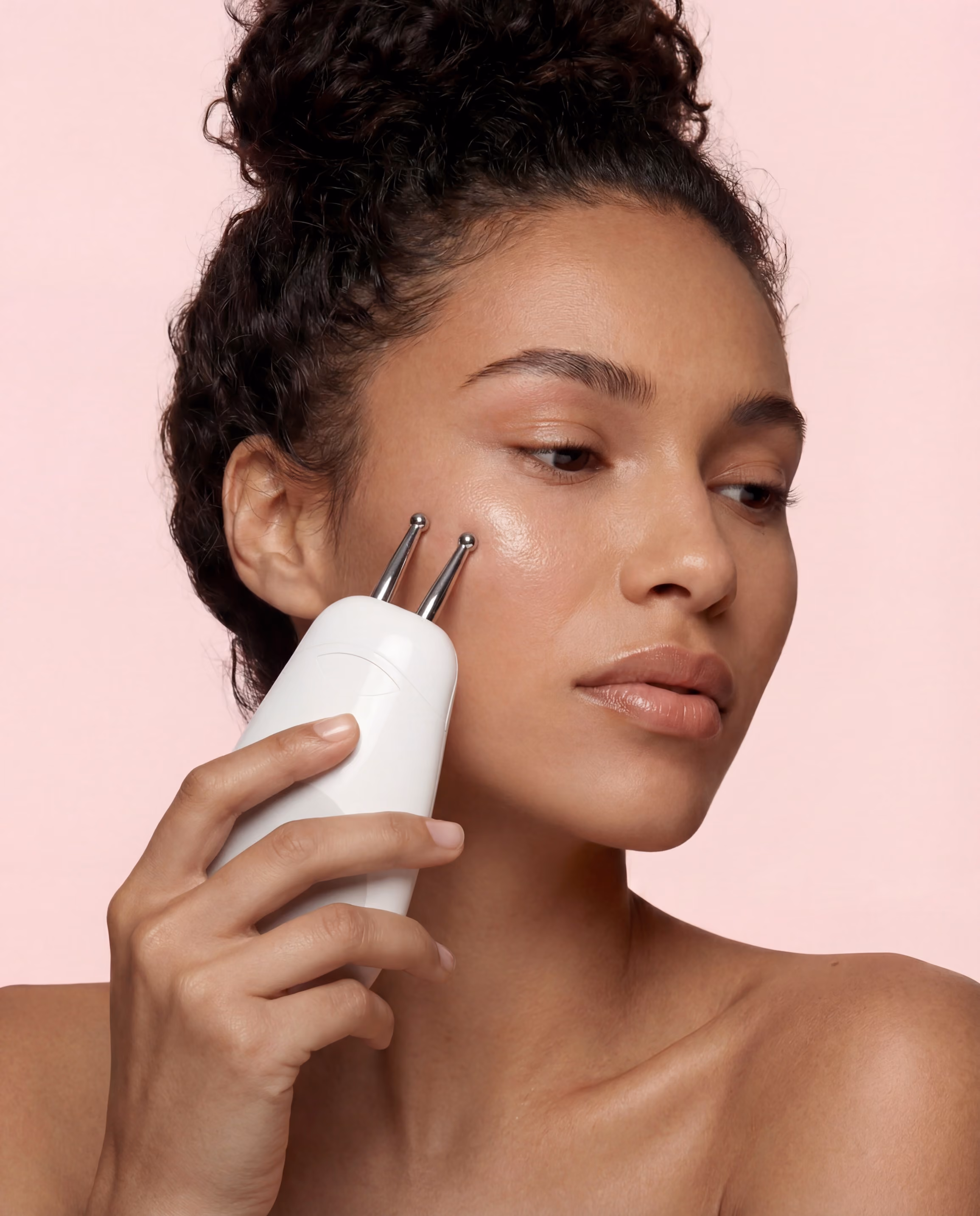 Woman with curly hair in a bun using a white facial toning device with two metal prongs on her cheek against a light pink background.