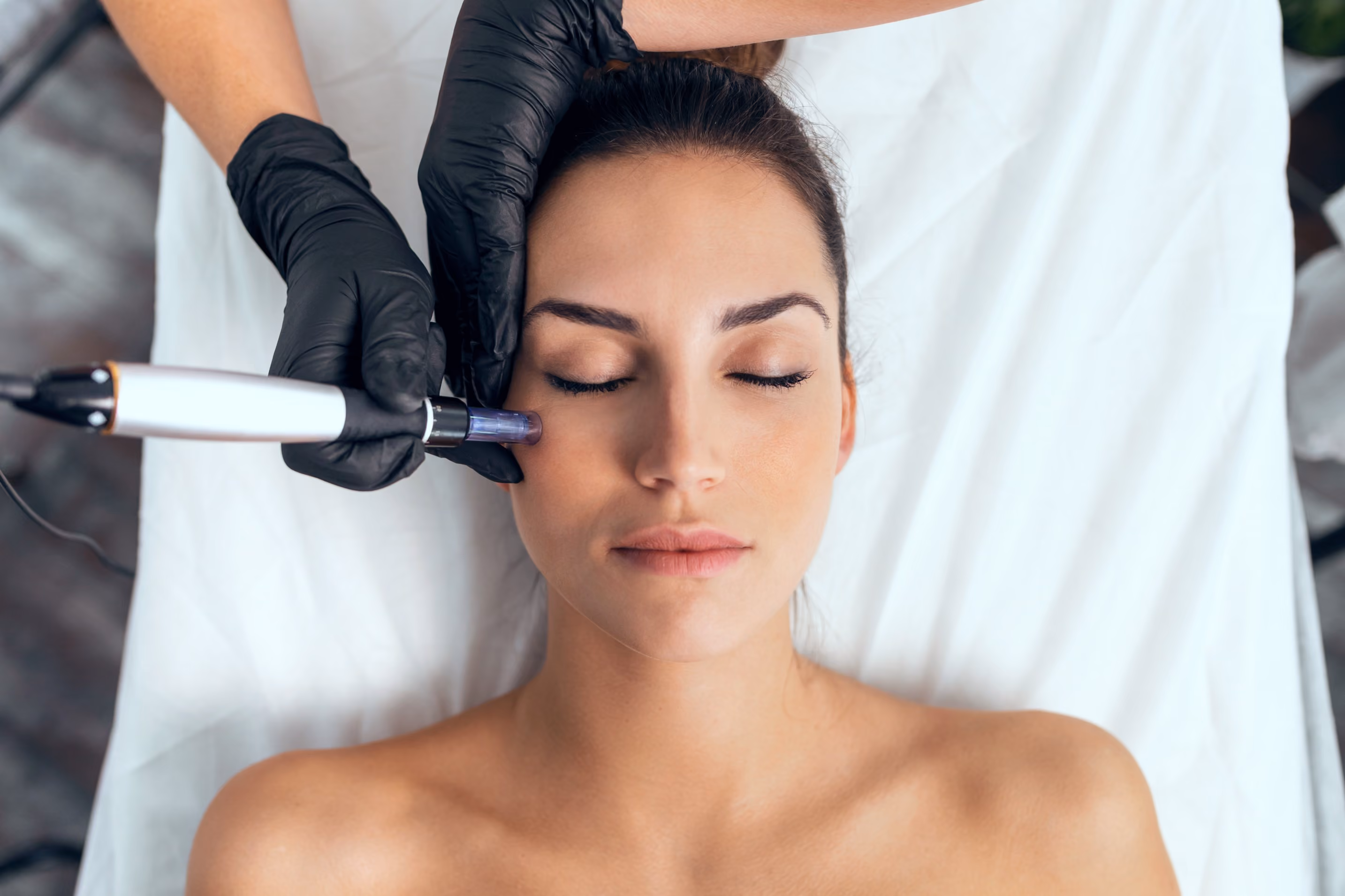 Close-up of a woman with eyes closed receiving a facial treatment from a person wearing black gloves using a handheld device on her cheek.