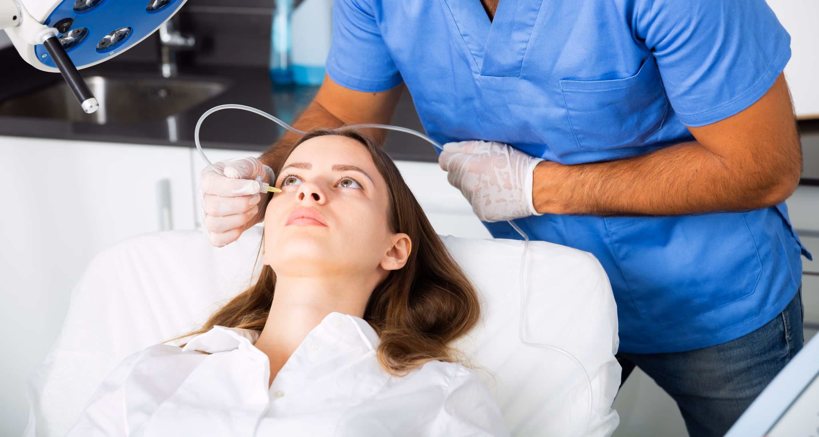 A medical professional in blue scrubs and gloves performs a facial treatment on a woman lying on a white chair, applying a tool near her nose in a clinical setting.