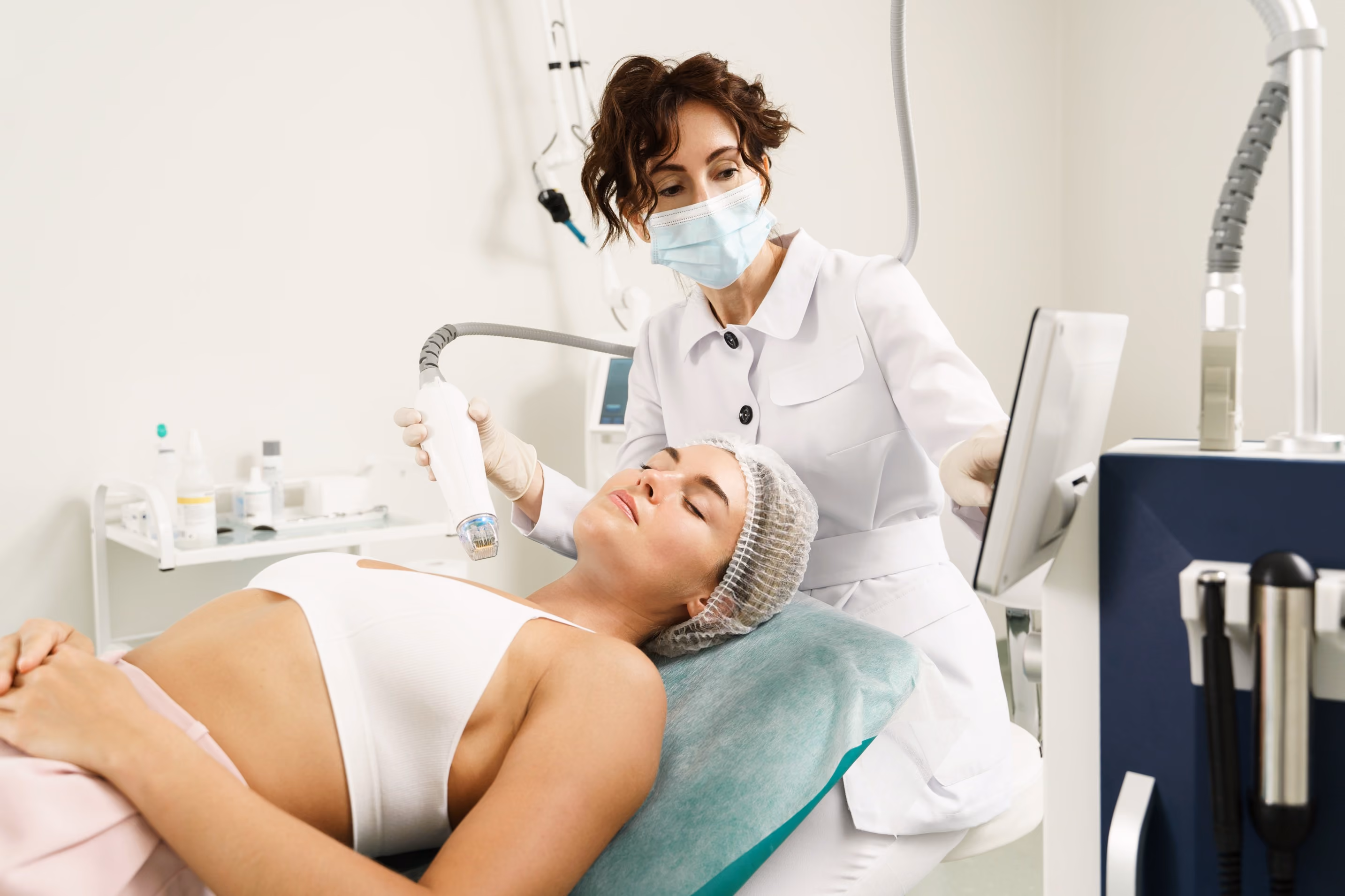 A woman lying on a medical bed receiving a facial treatment from a healthcare professional wearing a mask and gloves using a handheld device in a clinical setting.