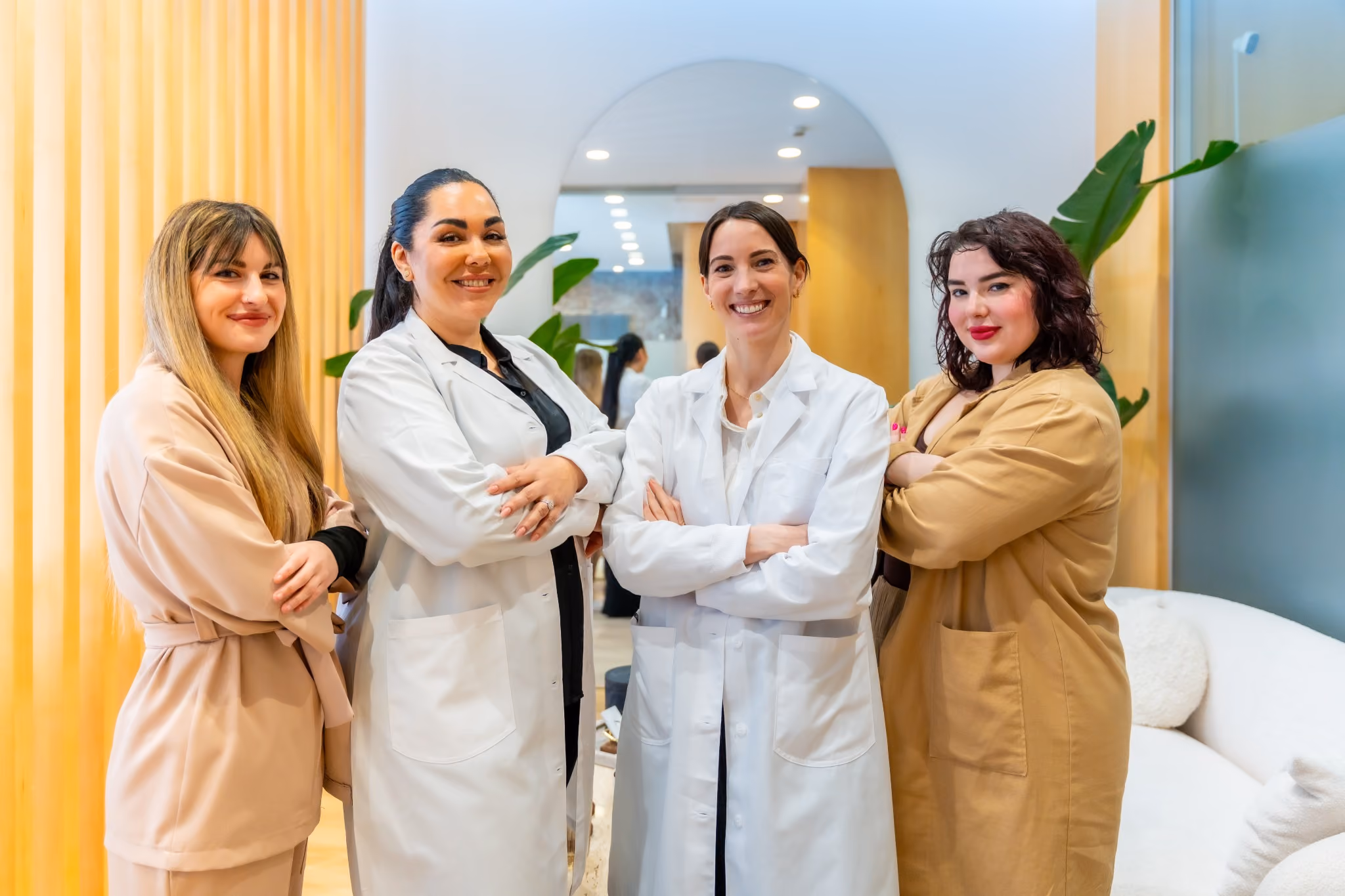 Four professional women standing indoors with arms crossed, two wearing white lab coats and two in beige outfits, smiling confidently in a modern office setting with plants and a round mirror in the background.