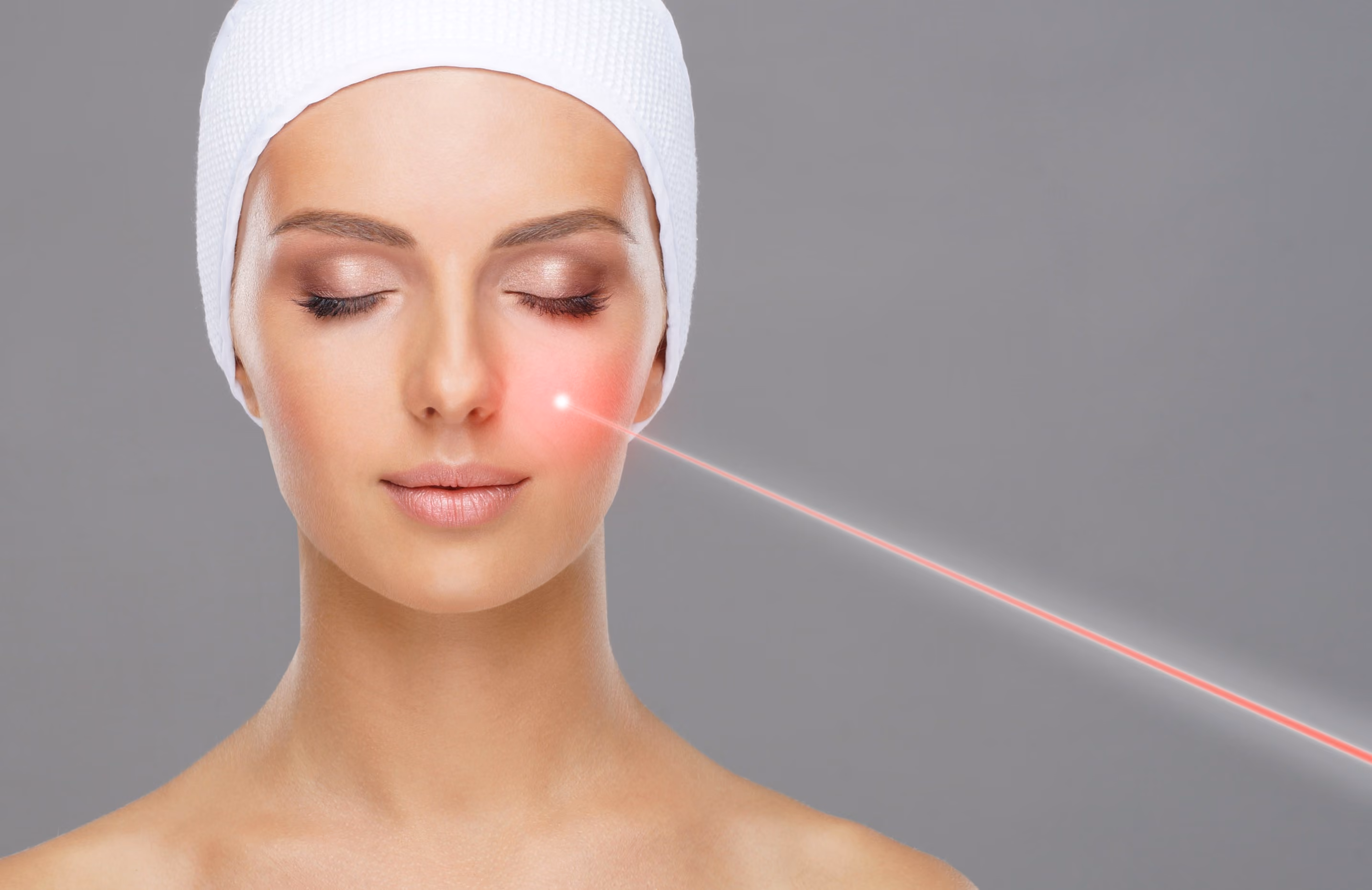 Woman with a white headband and closed eyes receiving a red laser treatment on her right cheek against a gray background.