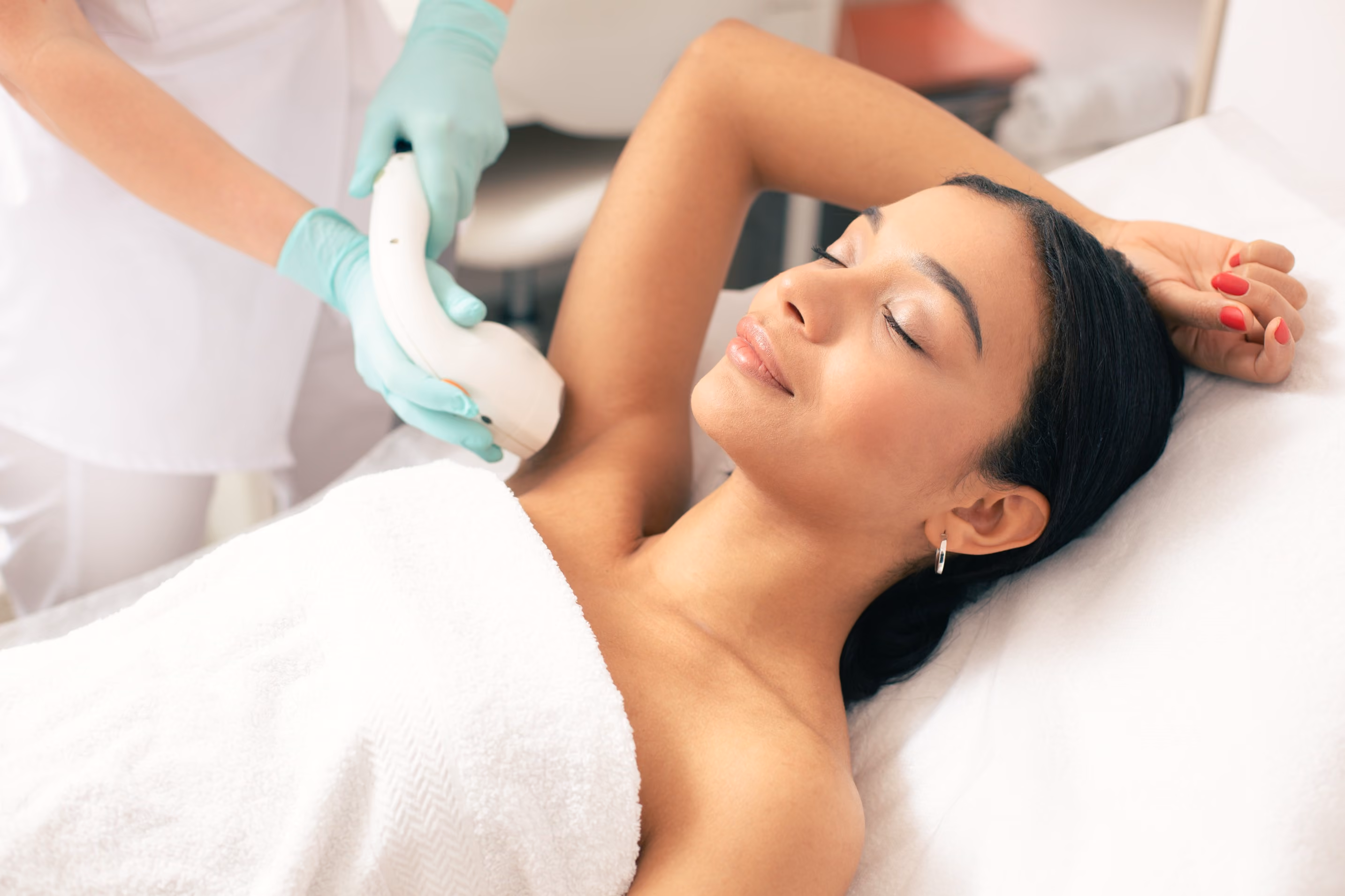 Woman lying on a spa bed with her eyes closed, receiving laser hair removal treatment on her underarm from a technician wearing green gloves.