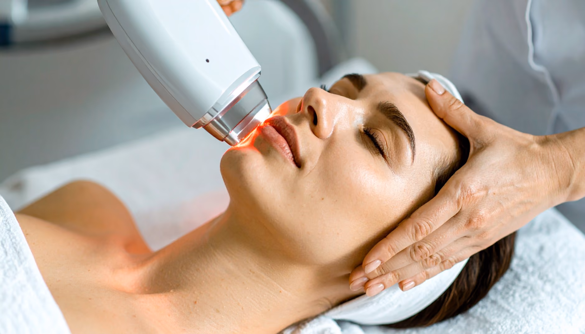Close-up of a woman receiving a facial laser treatment with a white handheld device emitting red light while a practitioner supports her head with one hand.