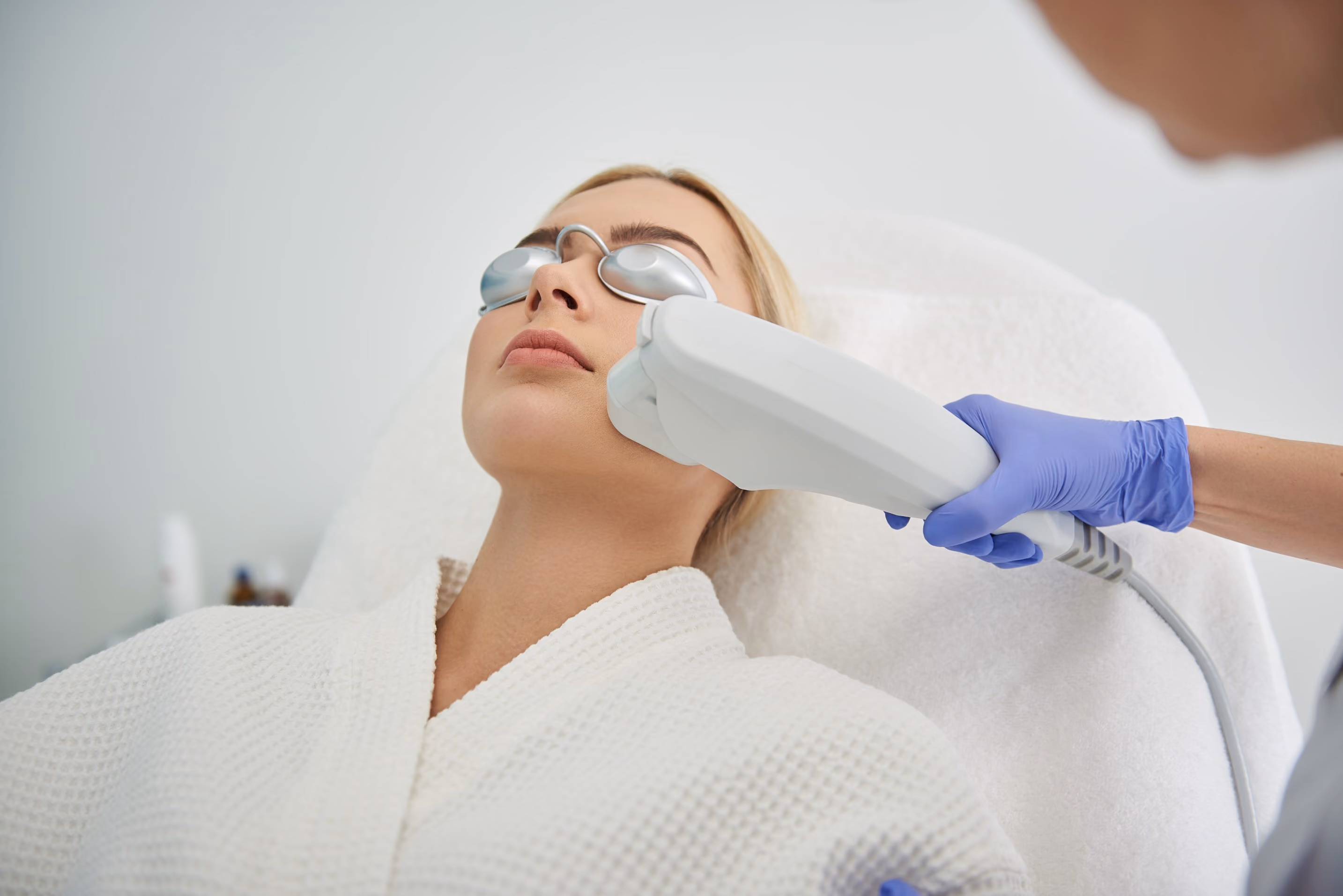 Woman wearing protective eyewear receiving a facial laser treatment from a professional wearing blue gloves, lying on a white treatment bed.