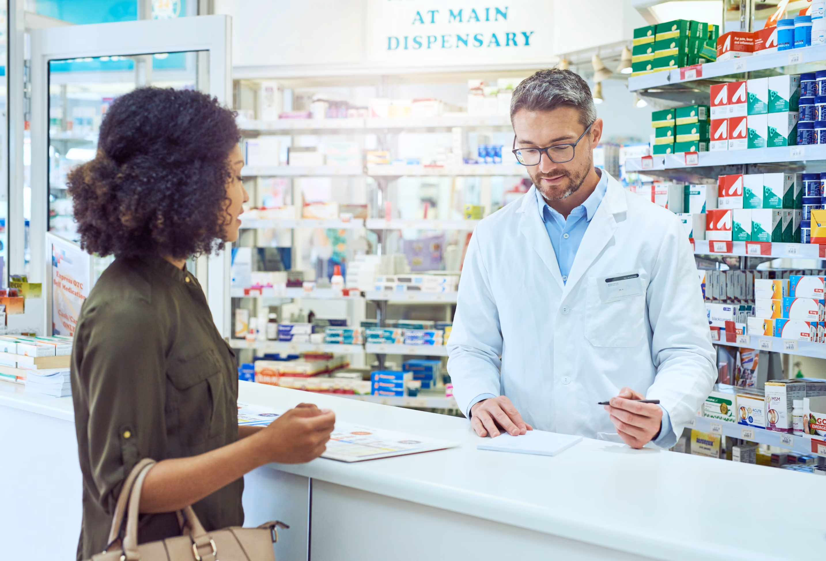 Pharmacist in a white coat consulting with a female customer at a pharmacy counter, with shelves of medication visible behind them.