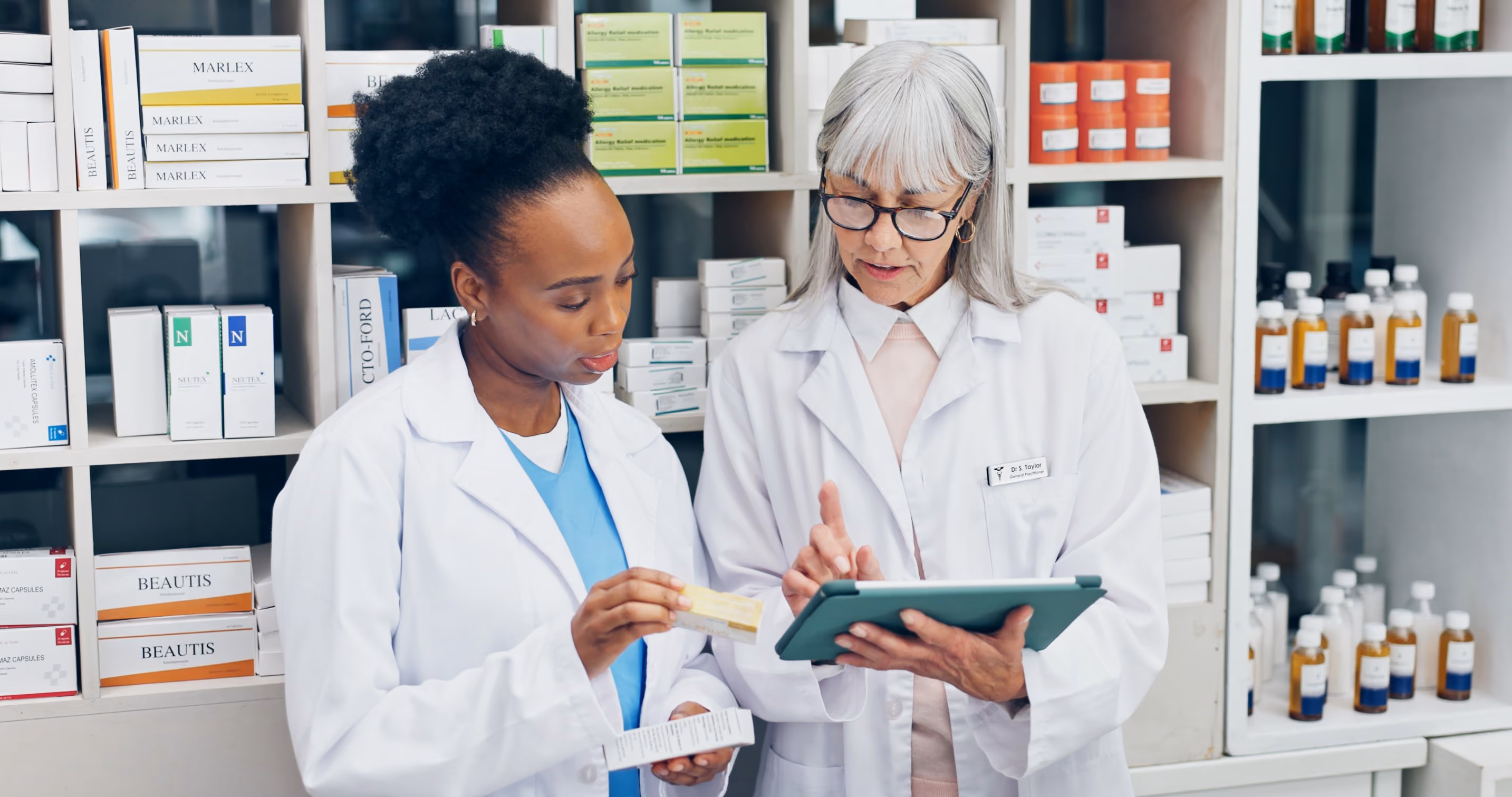 Two pharmacists in white coats discussing medication in a pharmacy; one holds a medicine box while the other points to a tablet screen.