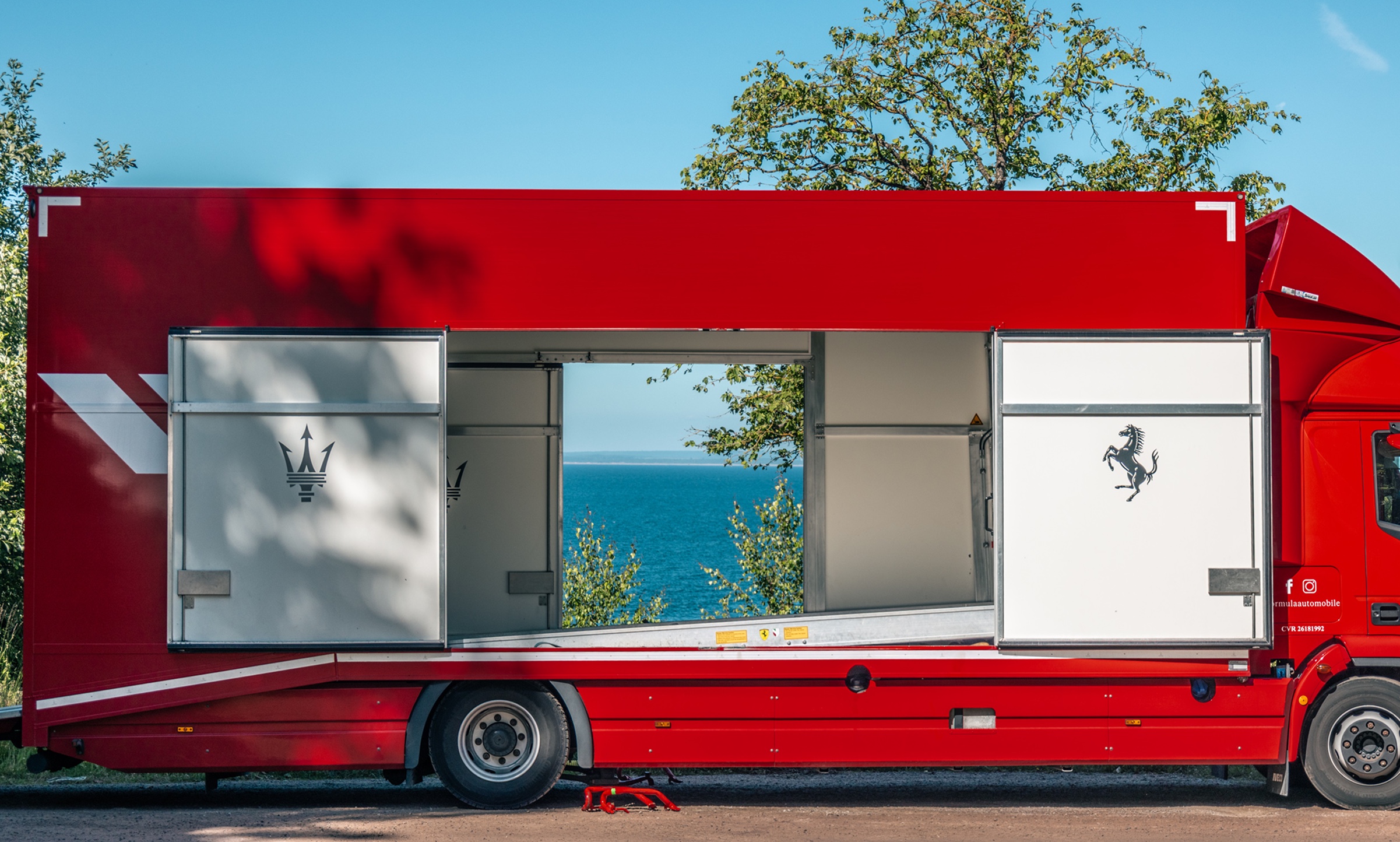 A red car transporter with open doors framing the sea