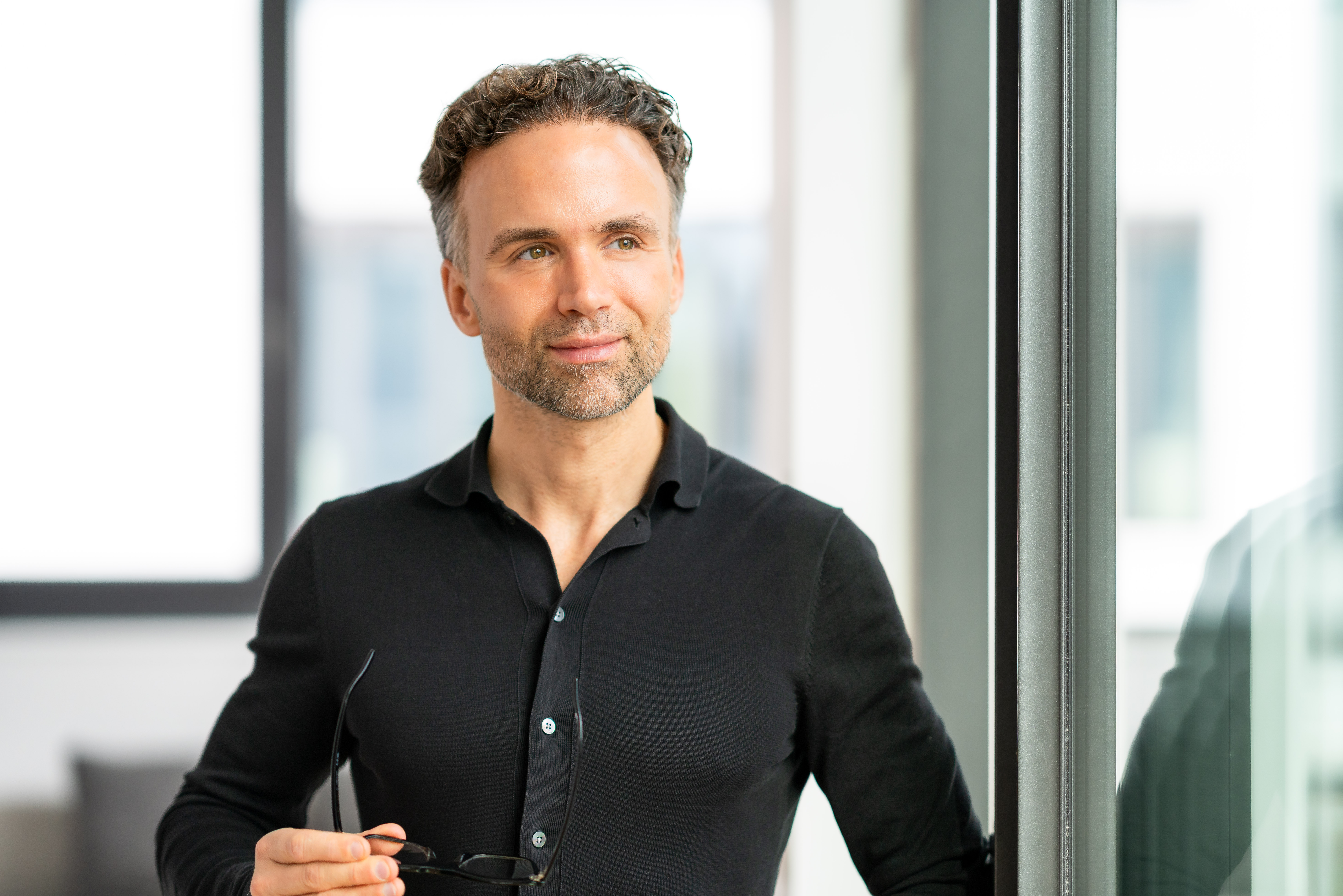 Smiling middle-aged man with short curly hair and light beard standing indoors holding glasses and looking thoughtfully out of a window.