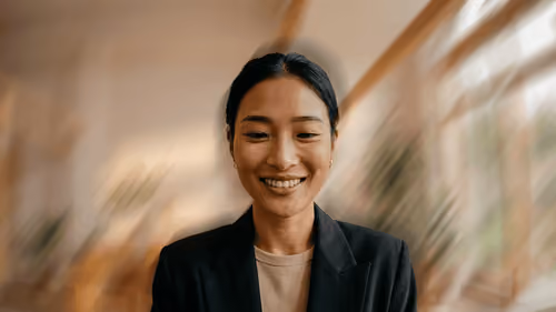 Smiling Asian woman with dark hair tied back, wearing a dark blazer and light shirt, in a softly blurred indoor setting with warm and natural light.